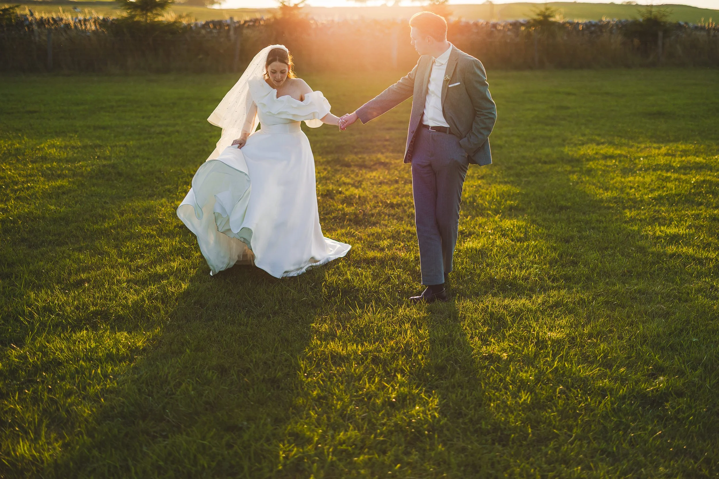 A bride and groom holding hands on a grassy field at sunset.