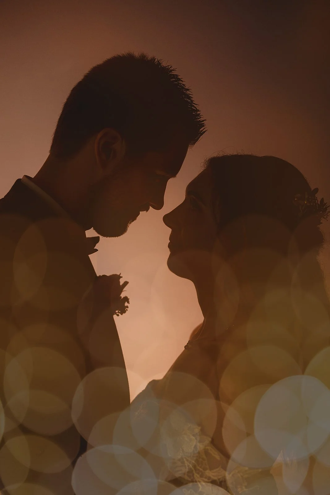 Silhouette of a couple gently touching foreheads in a romantic pose, with warm sunset lighting and double exposure bokeh effects in the foreground.