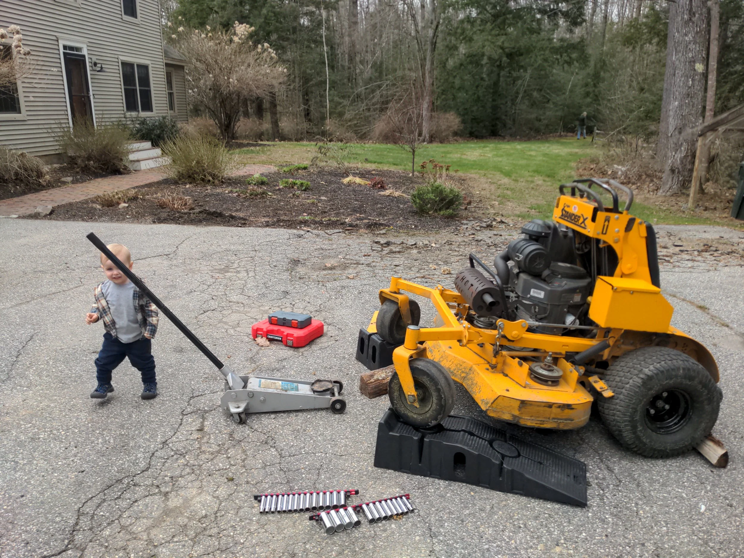 A toddler standing outside on cracked pavement, holding a long black pole, with tools and equipment around including a yellow riding mower, socket wrench set, and a red toolbox, with a house and garden in the background.