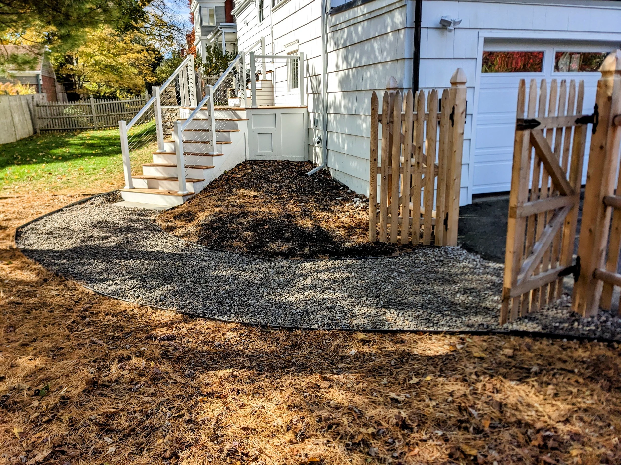 Backyard with newly installed steps and gravel pathway, wooden fence partially open, white house with garage in the background, and autumn trees with fallen leaves.