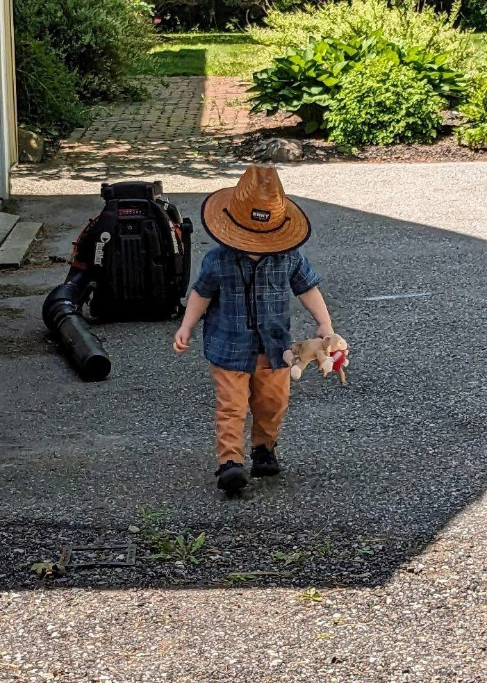 A young child walking outside on a driveway, wearing a large brown straw hat, a blue plaid shirt, orange pants, and black shoes, holding a toy teddy bear in one hand. There is a vacuum cleaner on the ground behind the child and some greenery in the background.