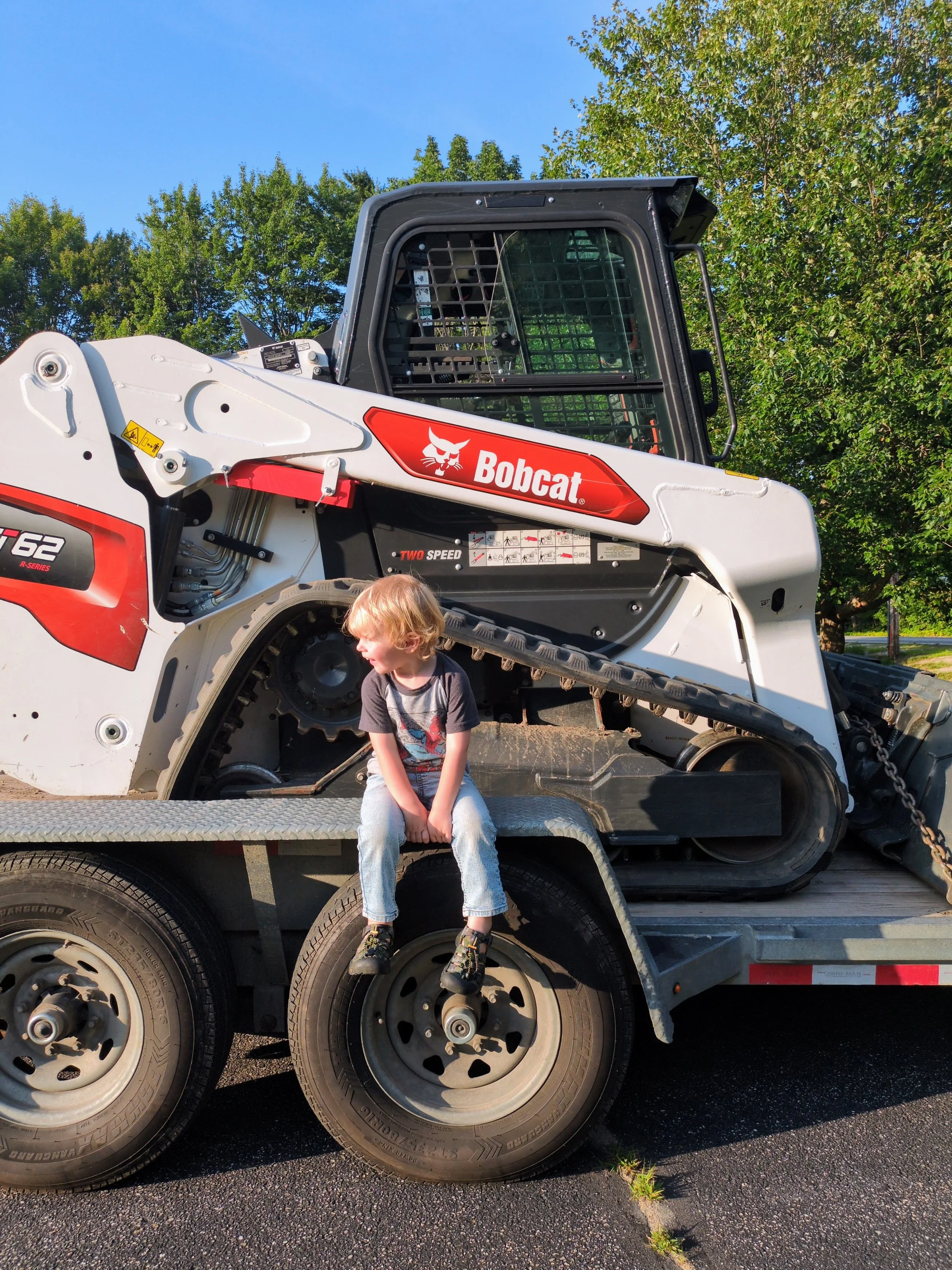 A young boy sitting on a flatbed trailer in front of a white Bobcat skid-steer loader, with trees and a blue sky in the background.