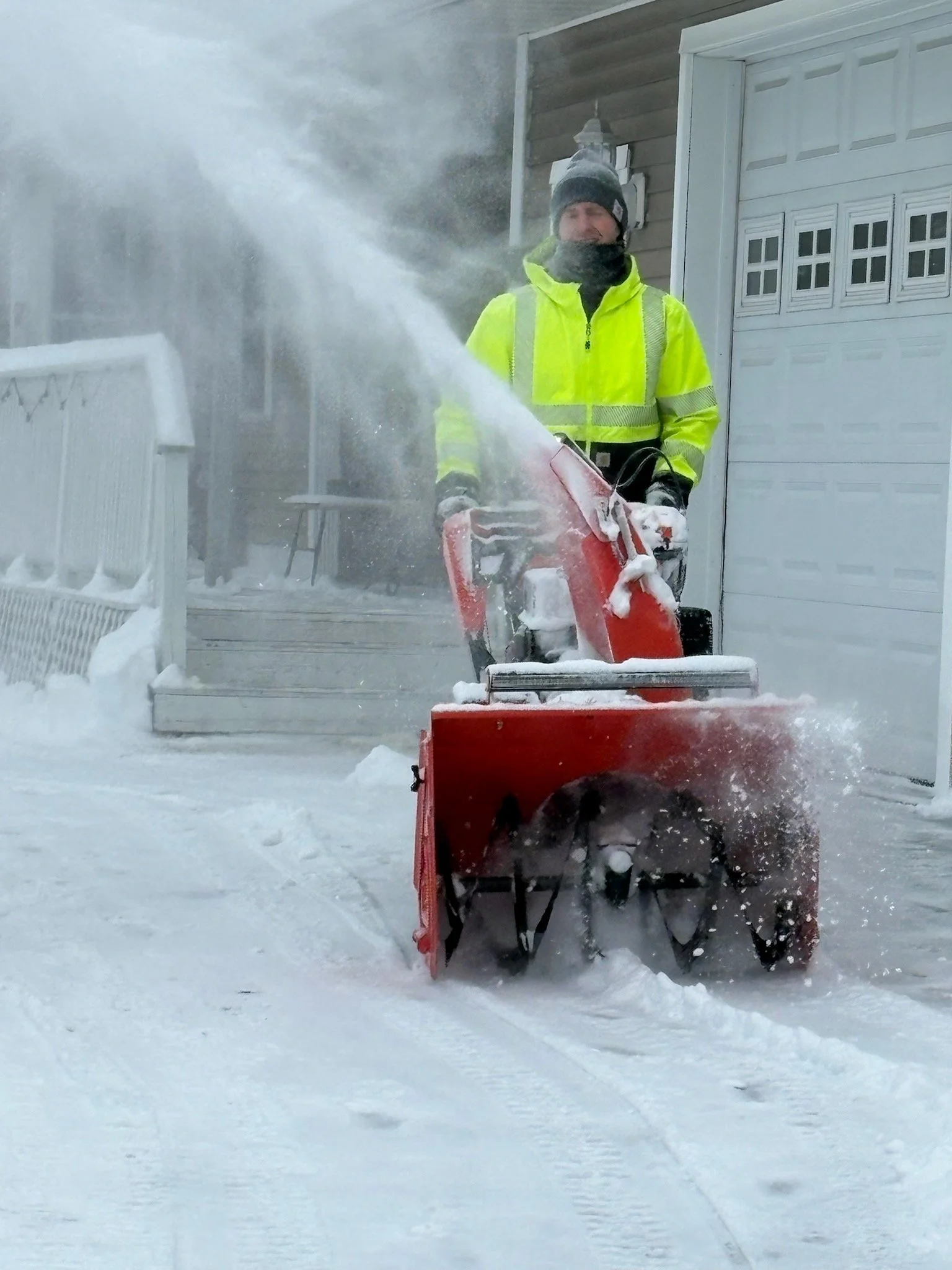 Person in a bright yellow winter jacket and gray hat clearing snow with a snowblower outside a house.
