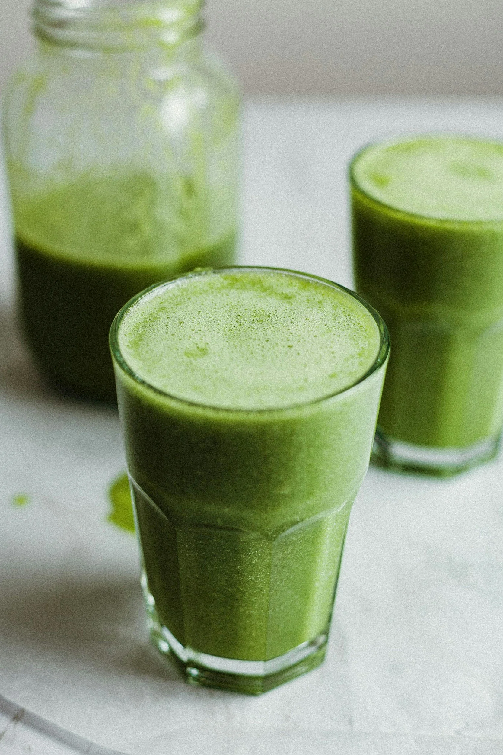Two glasses of bright green smoothie and a jar of green smoothie in the background.