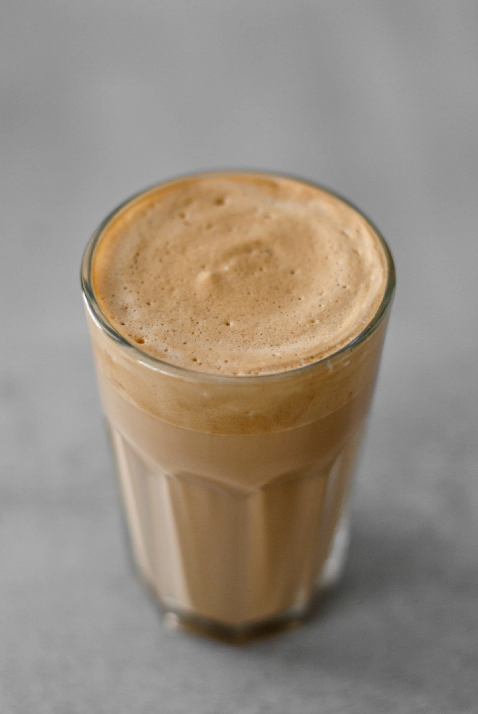 A tall glass of frothy iced coffee or latte with a creamy foam top on a gray surface.