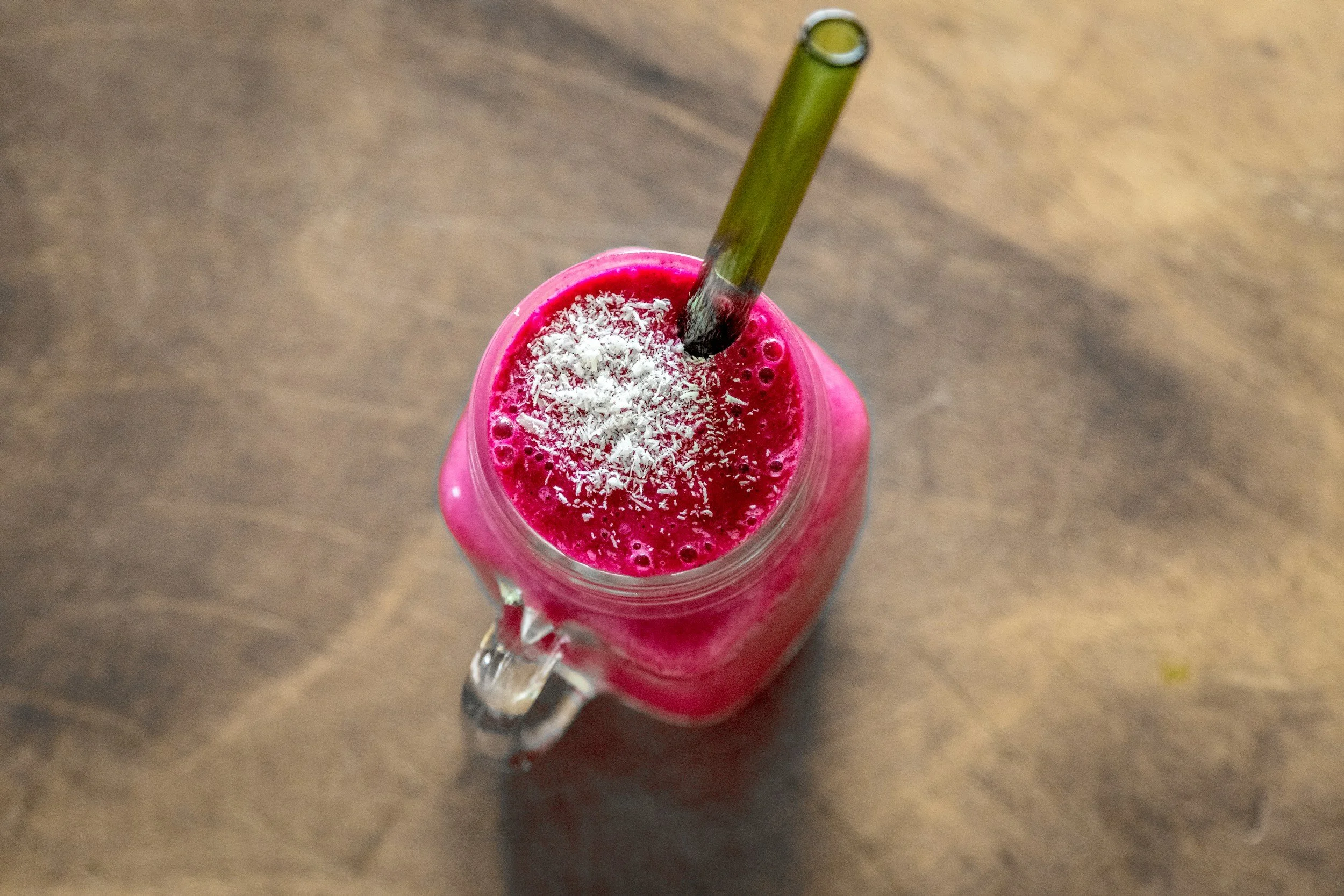 A pink smoothie in a glass jar with a metal straw, topped with shredded coconut, on a wooden surface.