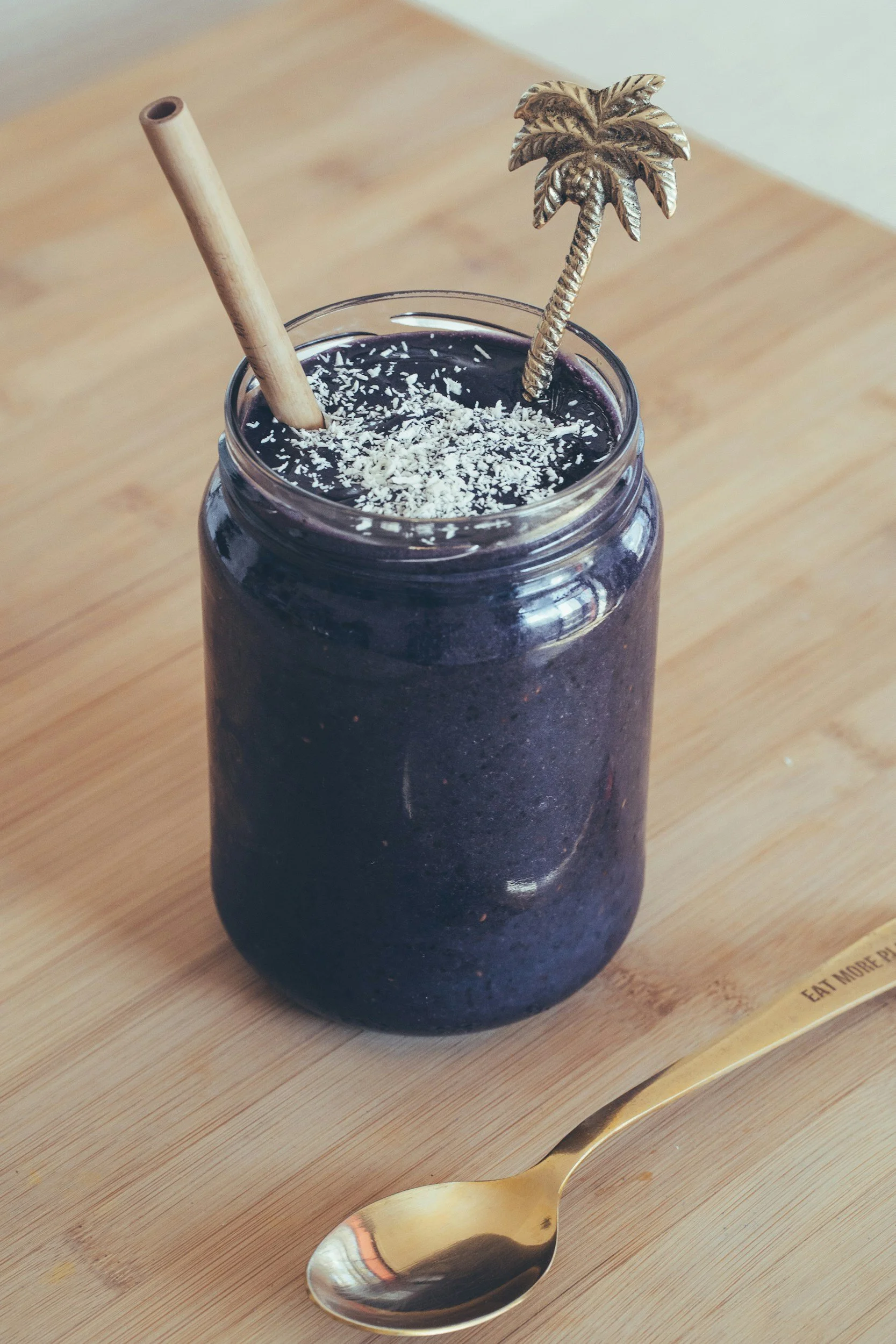 A dark purple or black smoothie in a mason jar topped with shredded coconut, with a bamboo straw and a gold palm tree stirrer. A gold spoon is on the wooden table beside the jar.