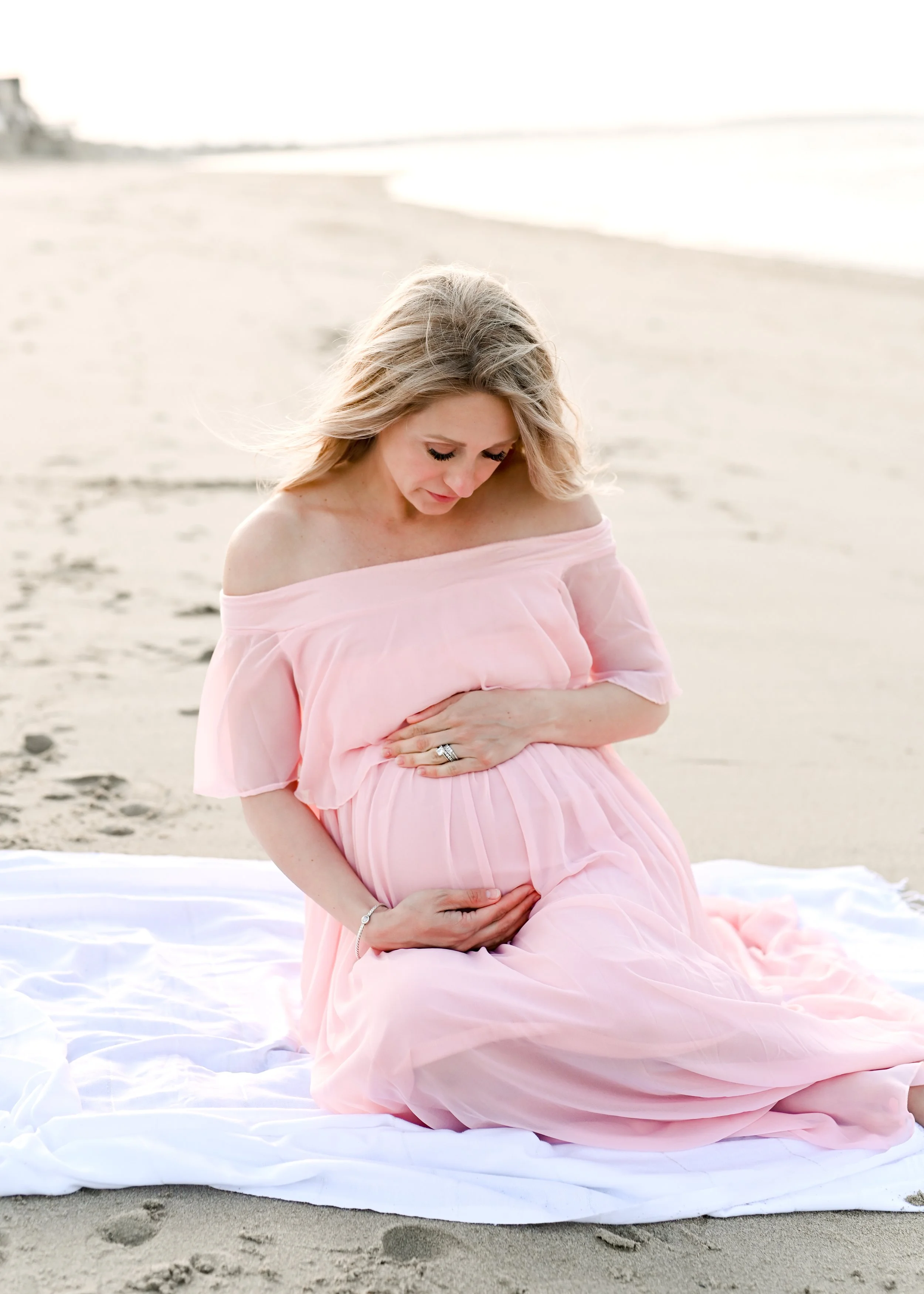 A pregnant woman in a pink off-the-shoulder dress sitting on a white blanket on the sandy beach, holding her belly and looking down.