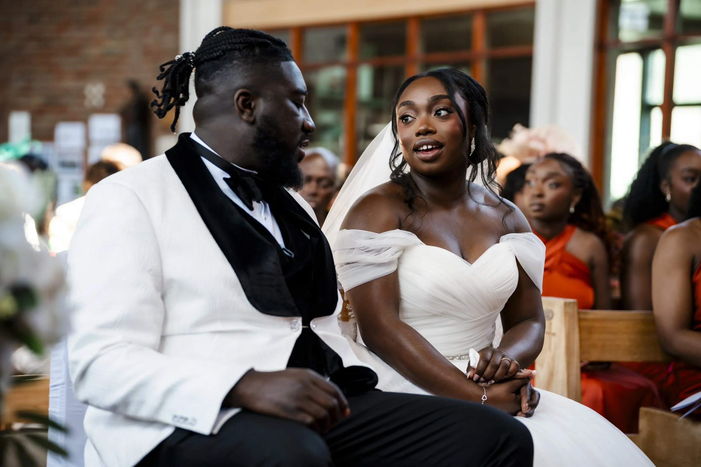 Bride and groom sitting together during their wedding ceremony inside a church, with wedding guests seated behind them.