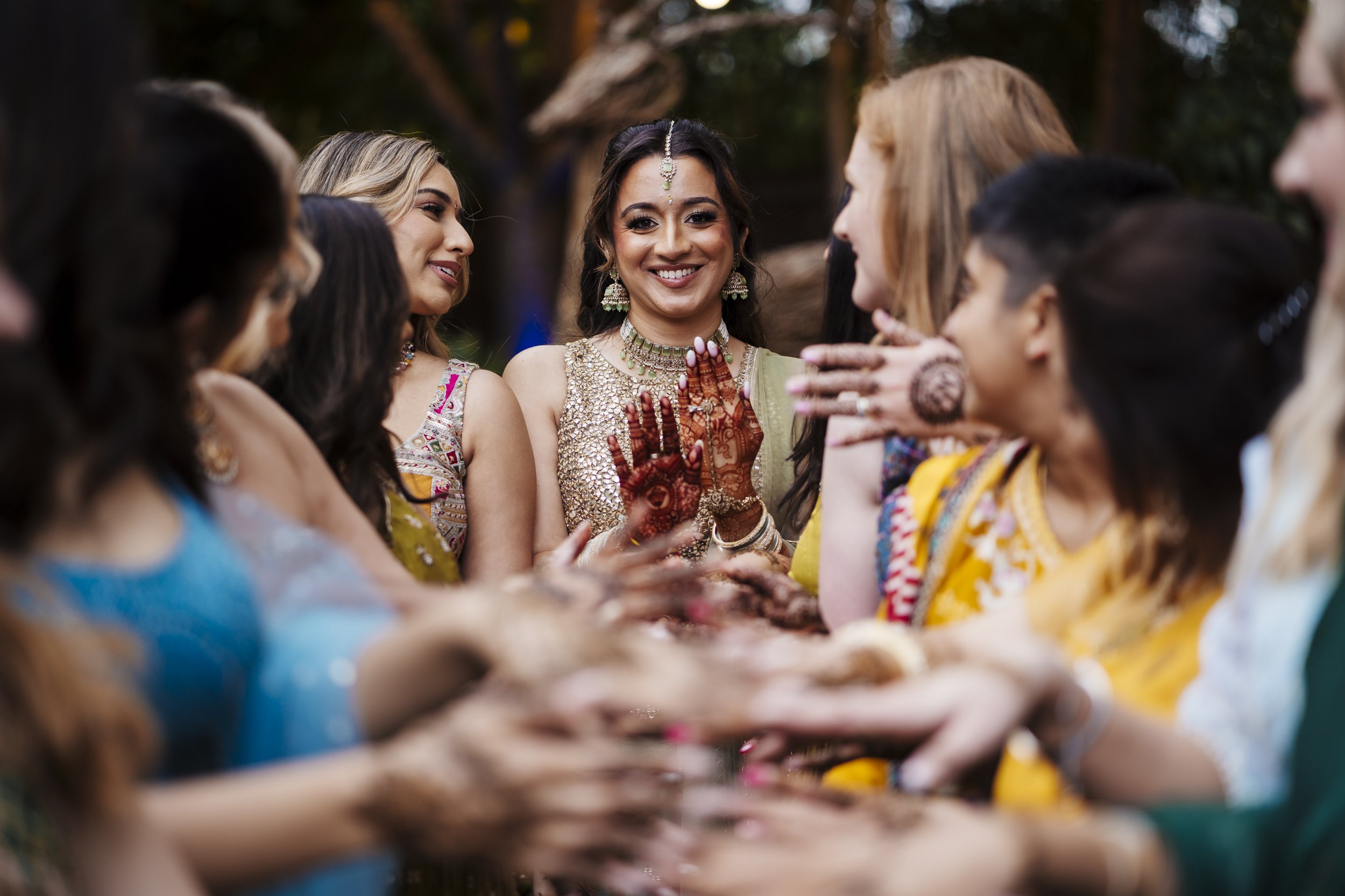 Group of women celebrating at an Indian wedding, wearing colorful traditional attire, with henna on their hands, smiling and talking.