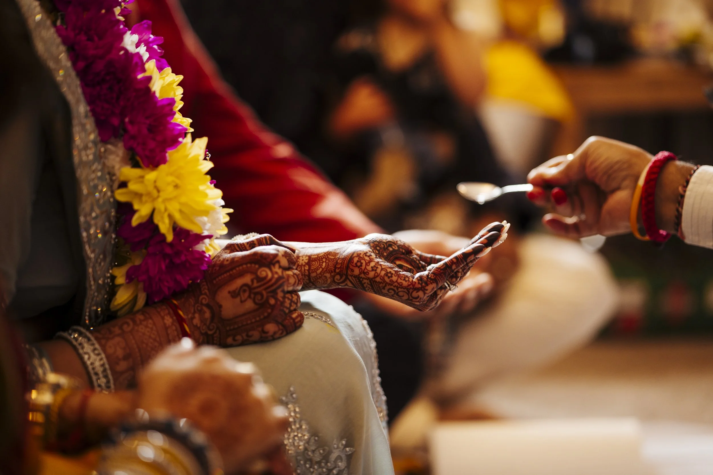 A person with ornate henna designs on their hand receives a tradition Indian rice blessing from another person during a wedding ceremony, decorated with yellow and purple flowers.