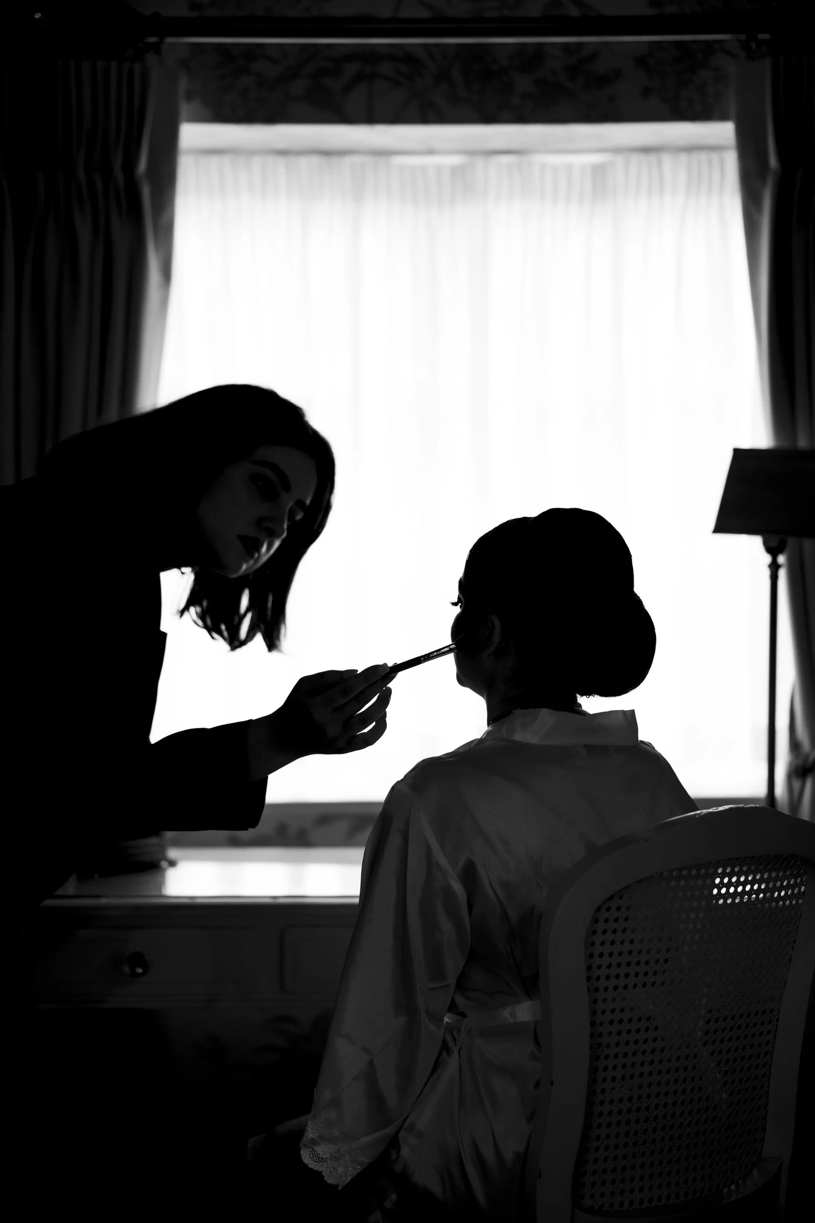 Silhouette of a woman applying makeup on another woman seated in front of a window with curtains.