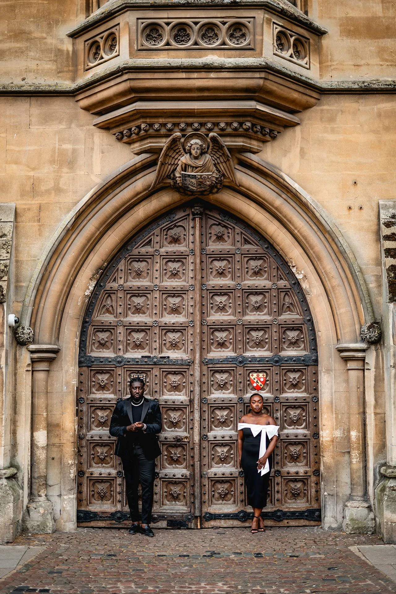 Two individuals standing in front of a large, ornate wooden door with medieval design, set in a stone building. The man on the left wears a black suit and holds a phone, while the woman on the right wears an off-the-shoulder black and white dress, an