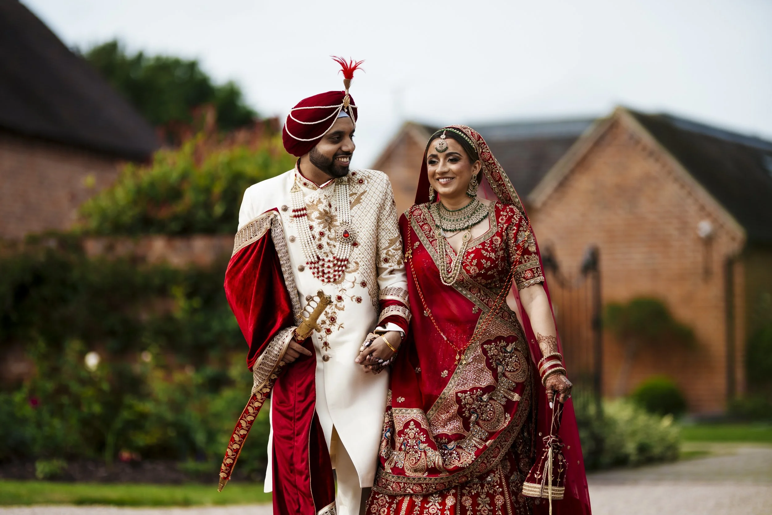 A bride and groom in traditional Indian wedding attire walking outdoors, smiling at each other.