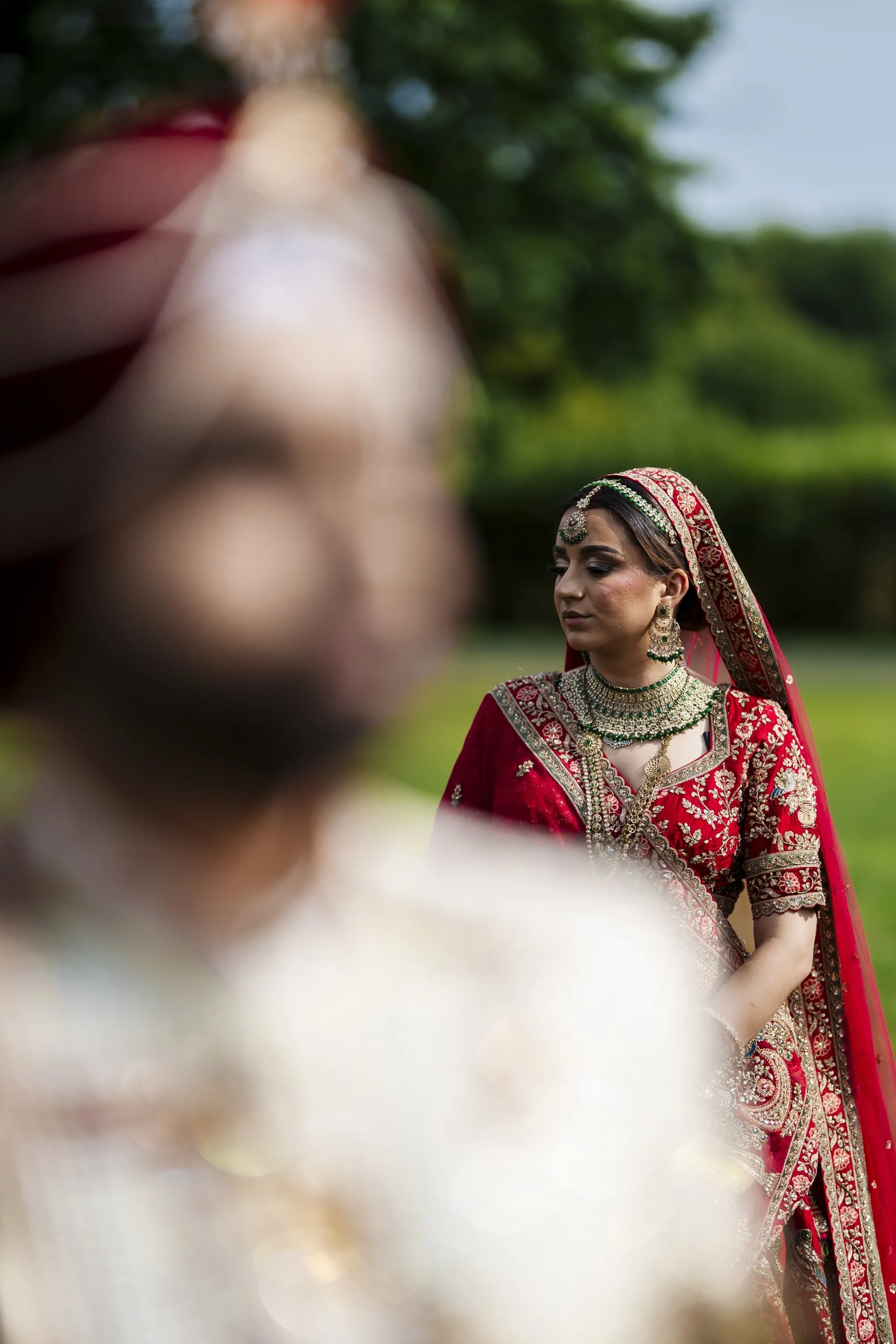 A woman dressed in traditional Indian wedding attire, including a red and gold embroidered sari, matching head covering, and heavy jewelry, stands outdoors with greenery in the background; a blurred figure in similar attire appears in the foreground.