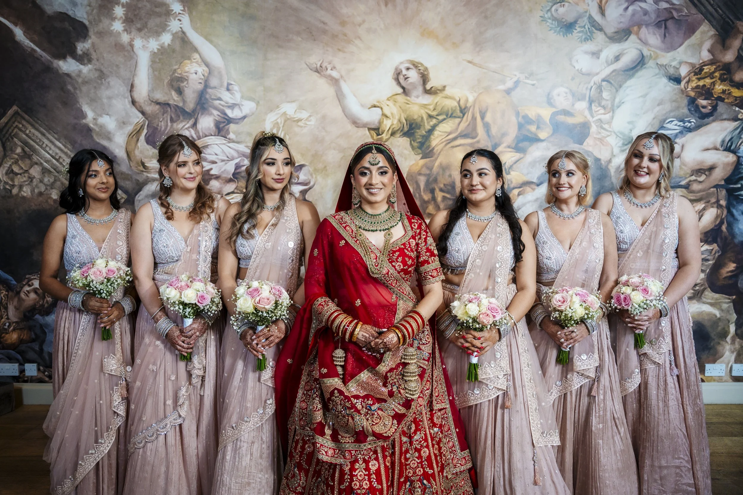A bride in a red and gold traditional Indian outfit standing with six bridesmaids in beige and silver sarees, all holding bouquets of pink and white roses, in front of a classical mural painting.