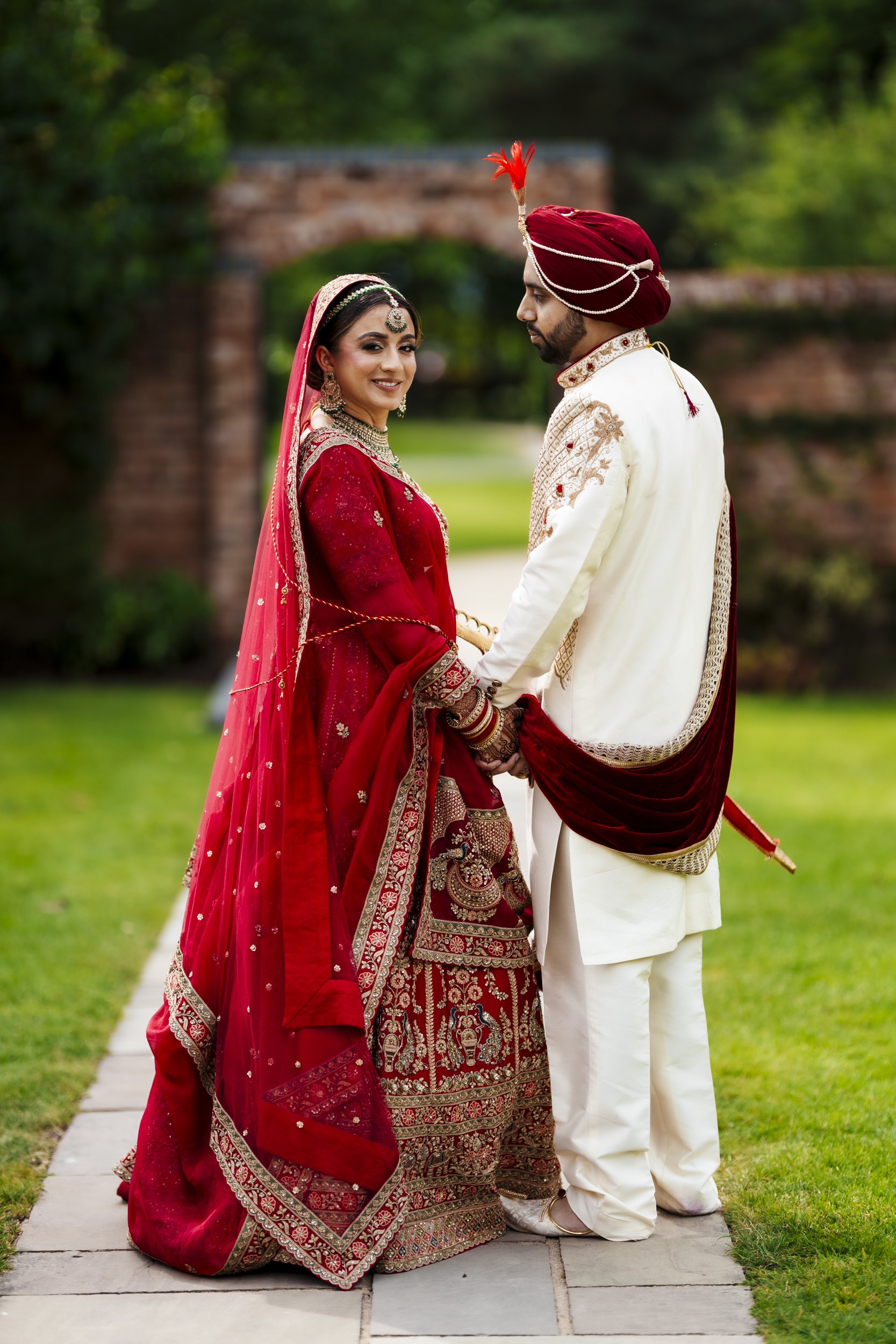 Hindu Punjabi, Gujrati, Indian bride and groom in traditional Indian wedding attire holding hands outdoors, with the bride smiling at the camera and the groom looking at her, standing on a stone pathway with green grass and trees in the background.