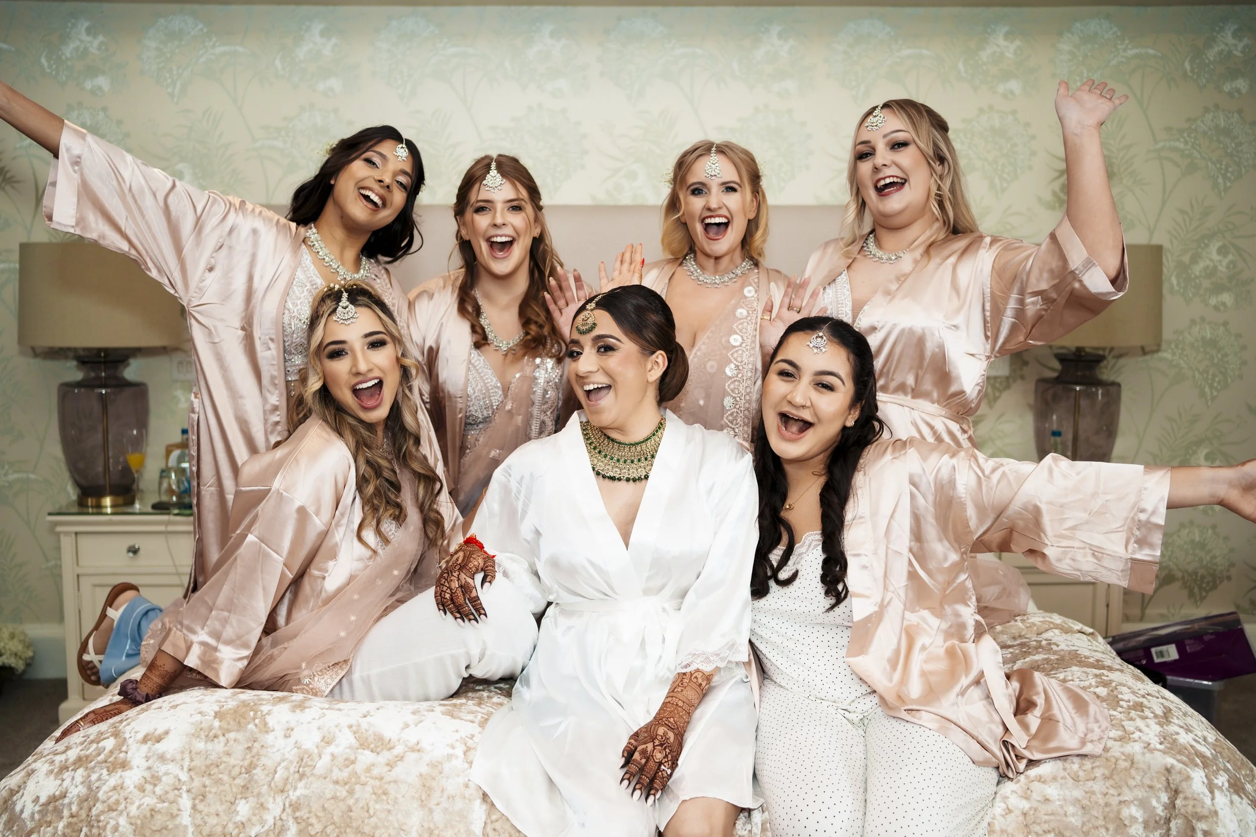 A group of women celebrating together, dressed in matching pink satin robes with jewelry, in a bedroom with floral wallpaper, smiling and making joyful gestures.