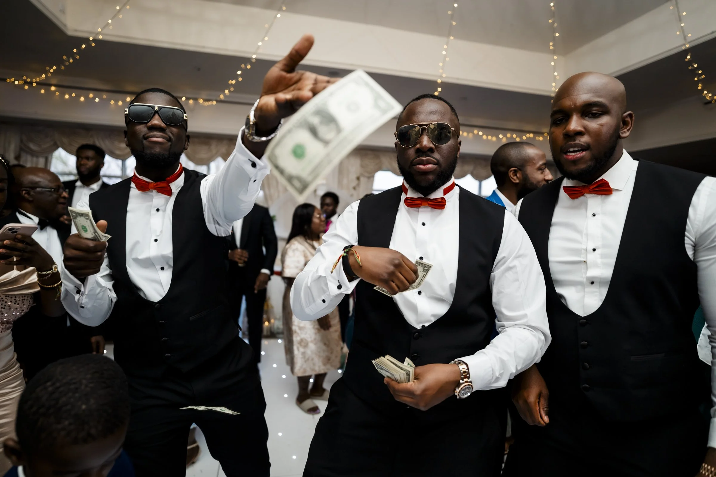 Three men dressed in black vests, white shirts, and red bow ties at a celebration, with one holding and throwing cash, some wearing sunglasses, in a decorated indoor venue.