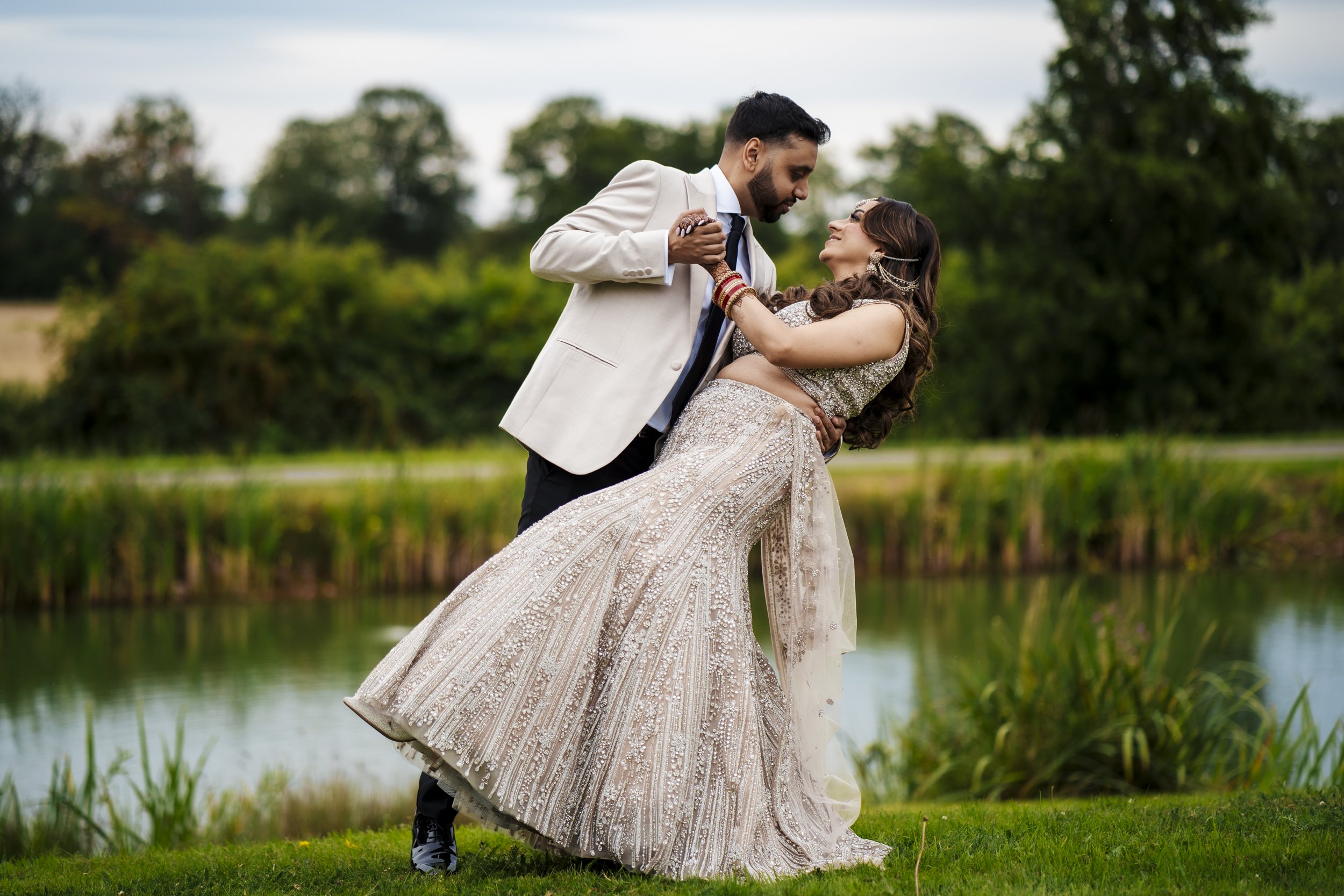 A couple dancing outdoors near a pond, the man holding the woman as she leans back in a gown and a man in a beige suit and tie, set against trees and a cloudy sky.