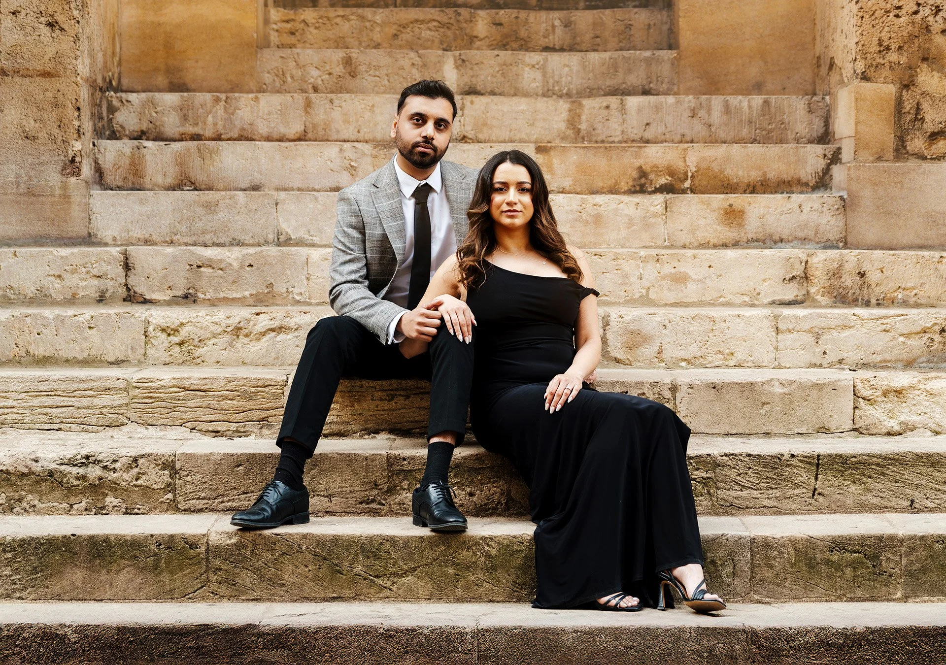 A man and woman sit on stone steps outdoors. The man wears a gray plaid blazer, white shirt, black tie, black pants, and black shoes. The woman wears a black off-the-shoulder dress and high heels. The man has his hand on the woman's arm, and both loo
