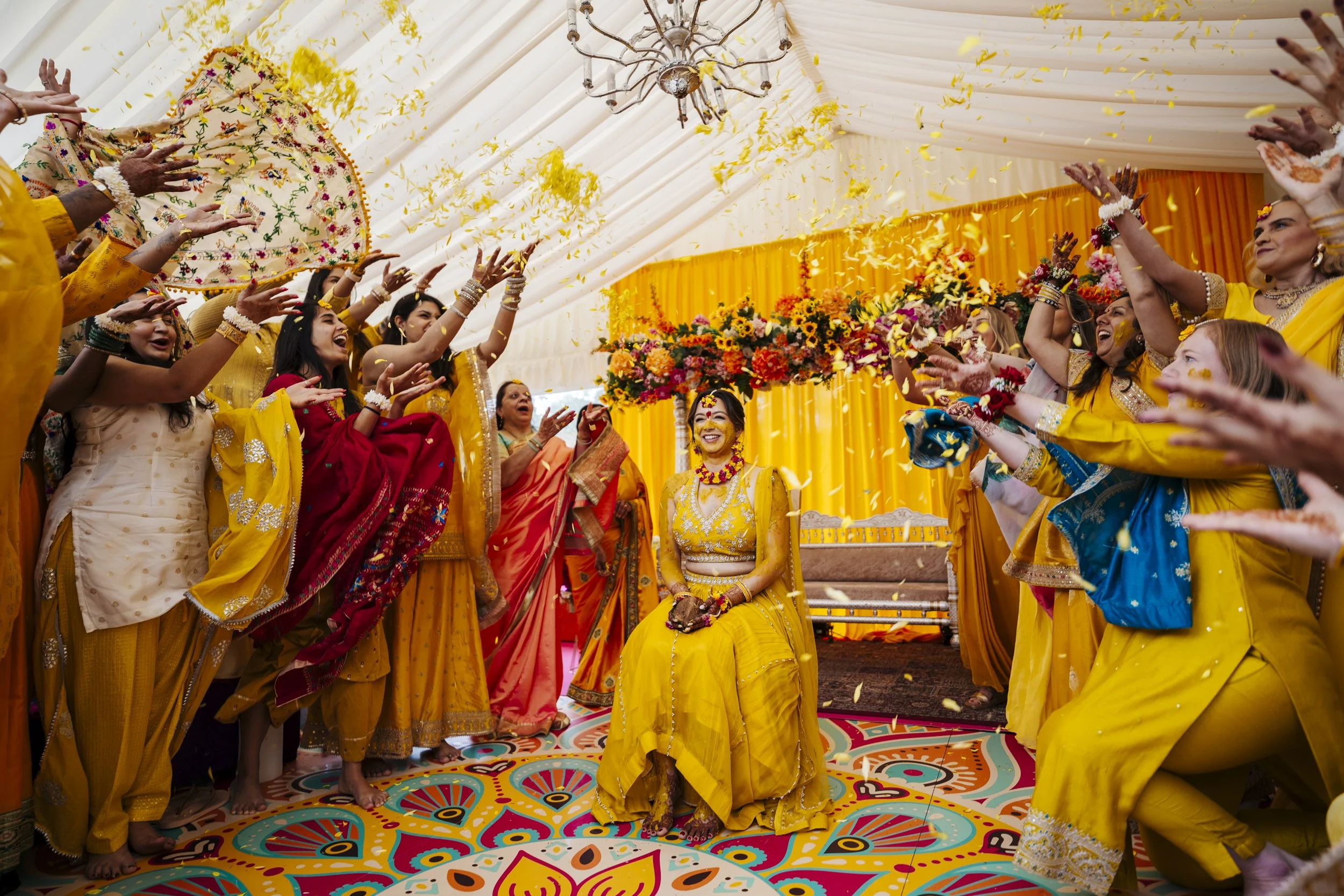 Indian bride dressed in yellow, sitting on a chair, surrounded by women throwing flower petals during a wedding ceremony in a decorated tent.