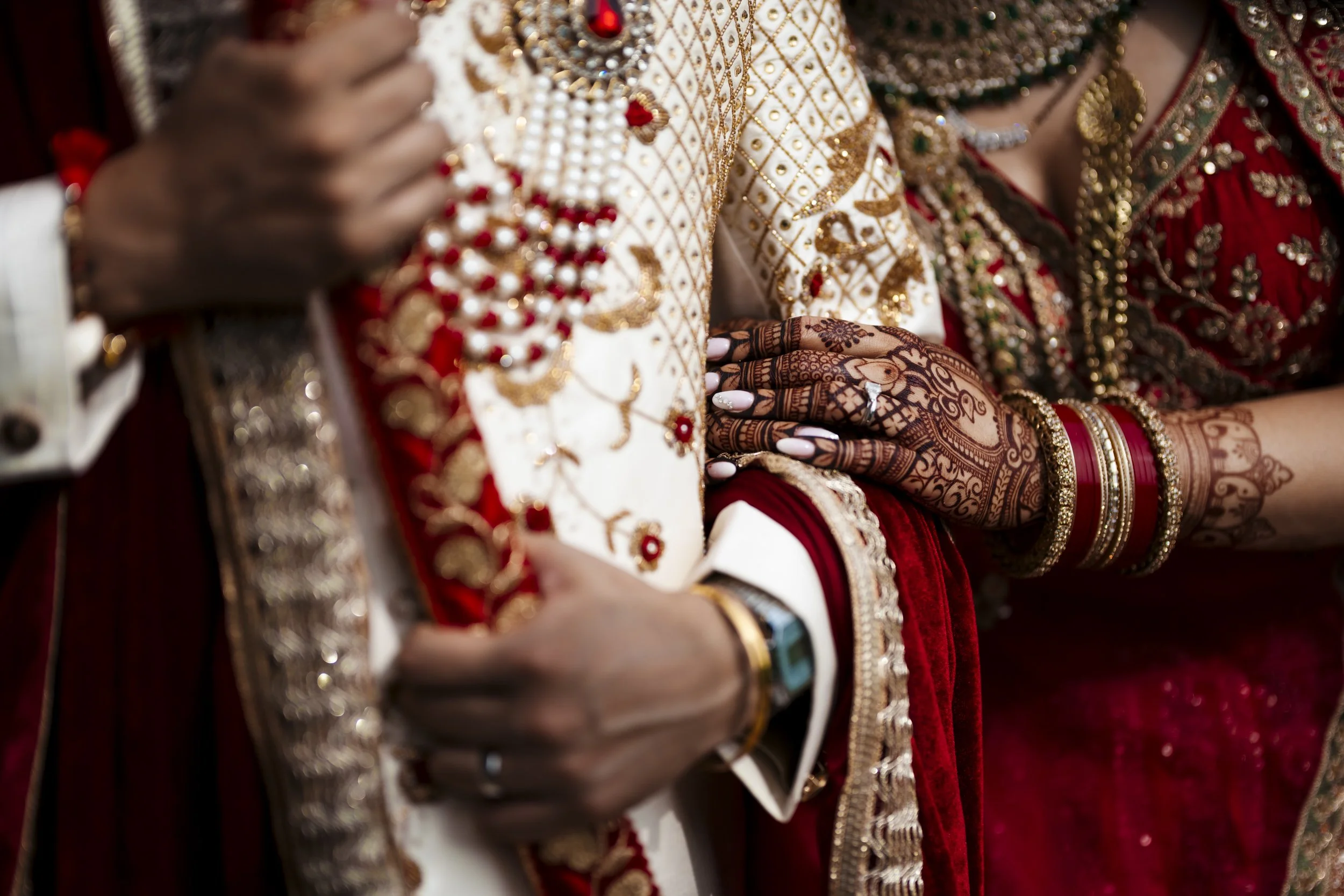 Close-up of a bride and groom's hands during a traditional Indian wedding. The bride's hands are decorated with intricate henna designs and various bangles, resting on the groom's arm. The groom is dressed in a richly embroidered sherwani with gold, 
