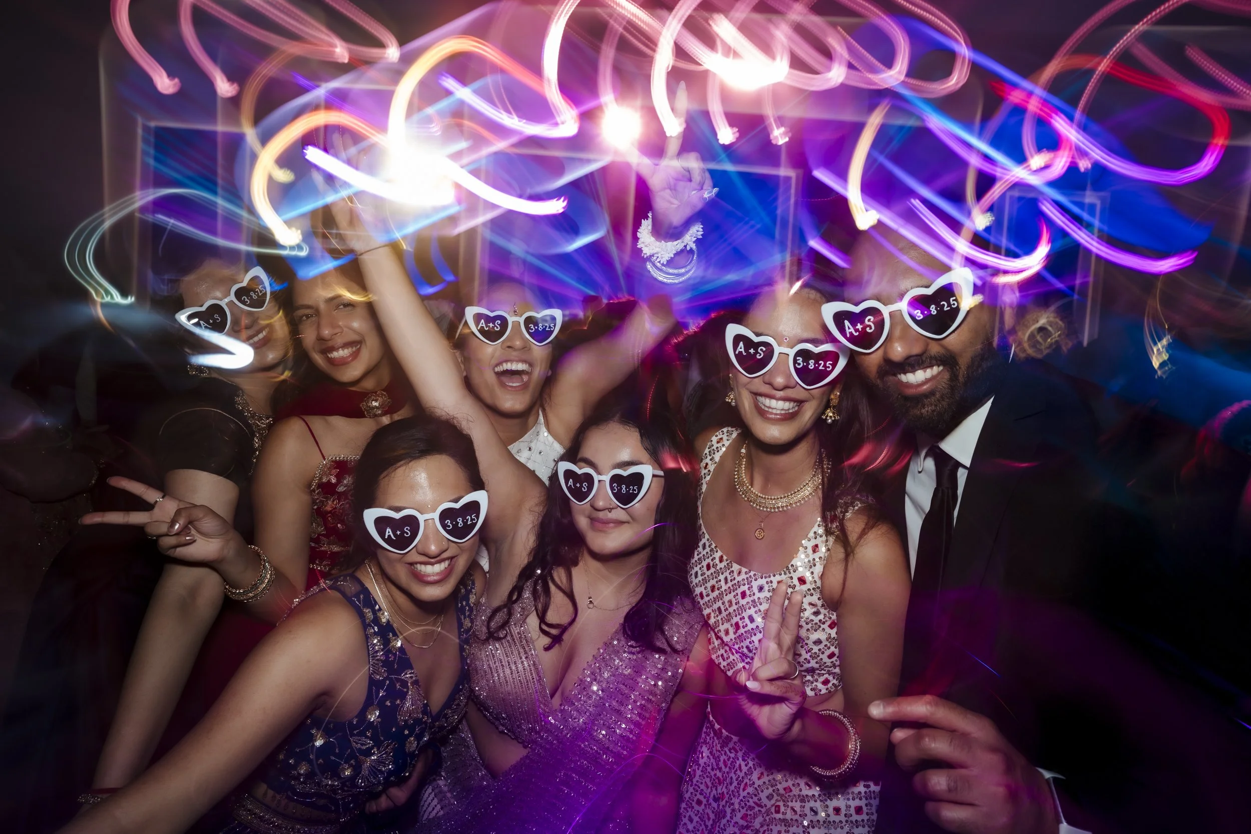 Group of friends celebrating at a party, wearing sunglasses with vows and date written on them, surrounded by colorful light trails.