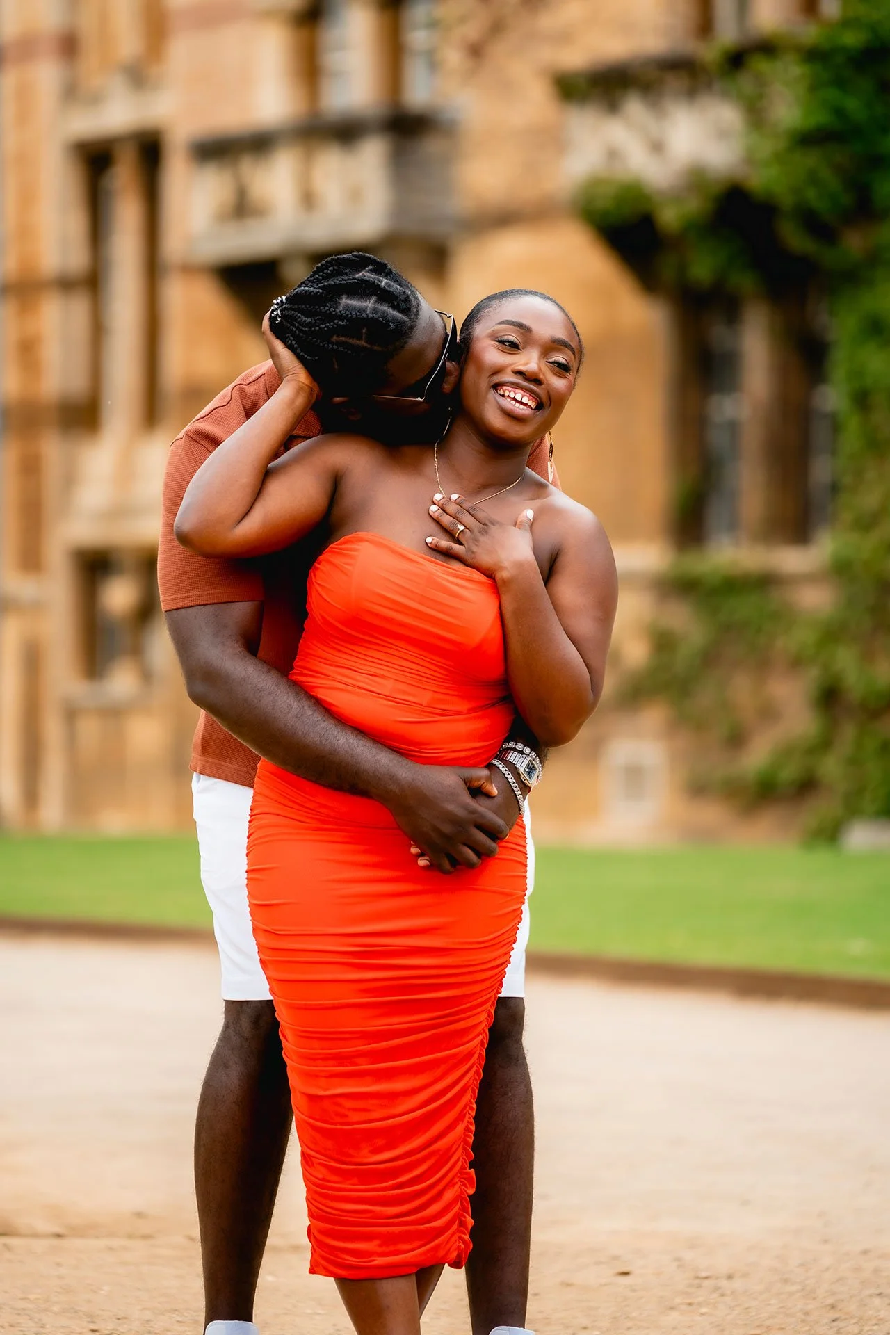 A happy couple embracing outdoors, woman in an orange dress and man in a brown shirt and white shorts, with a historic brick building in the background.