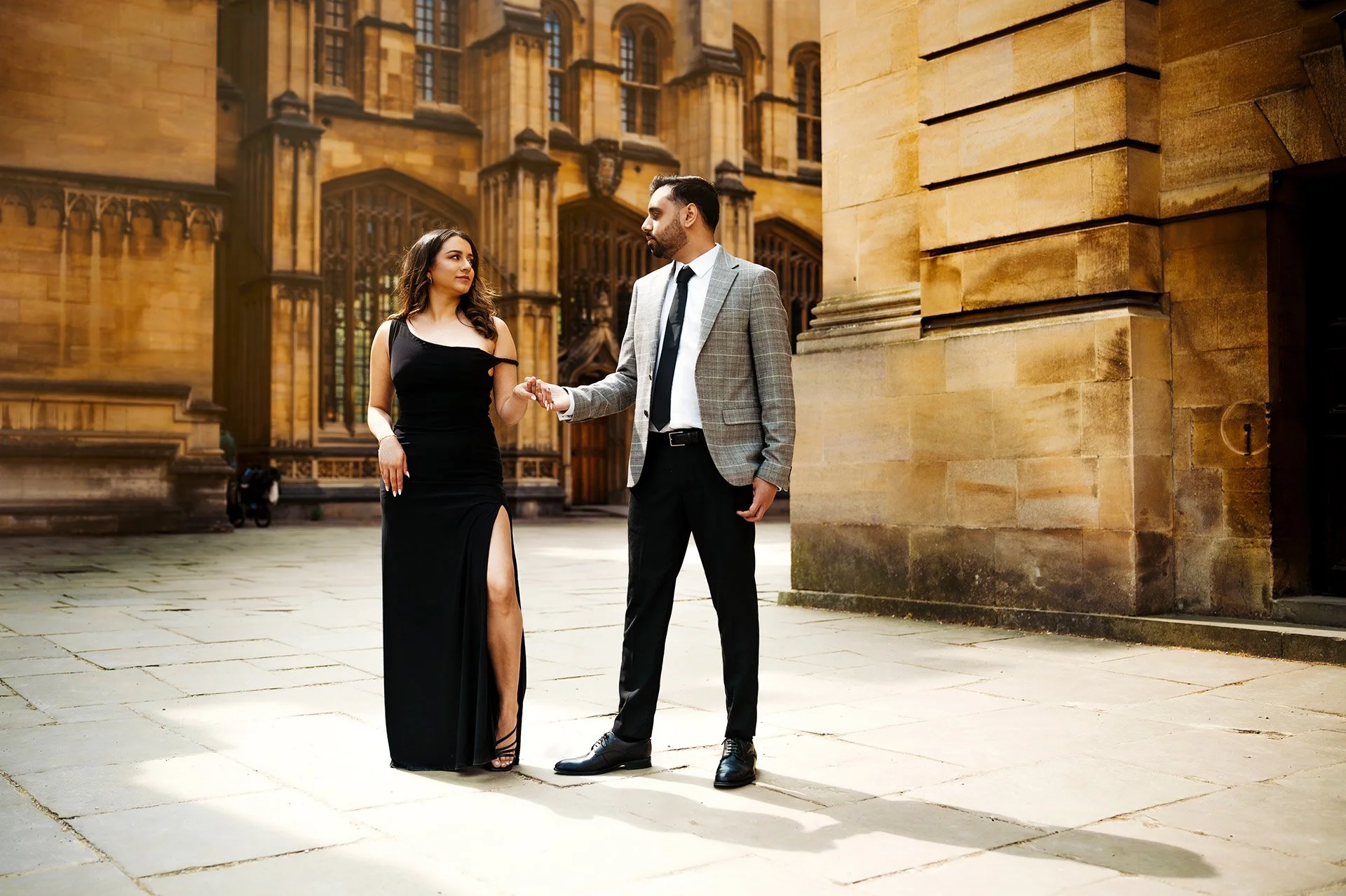 A man and a woman are holding hands and walking outside a historic stone building, dressed in formal attire.