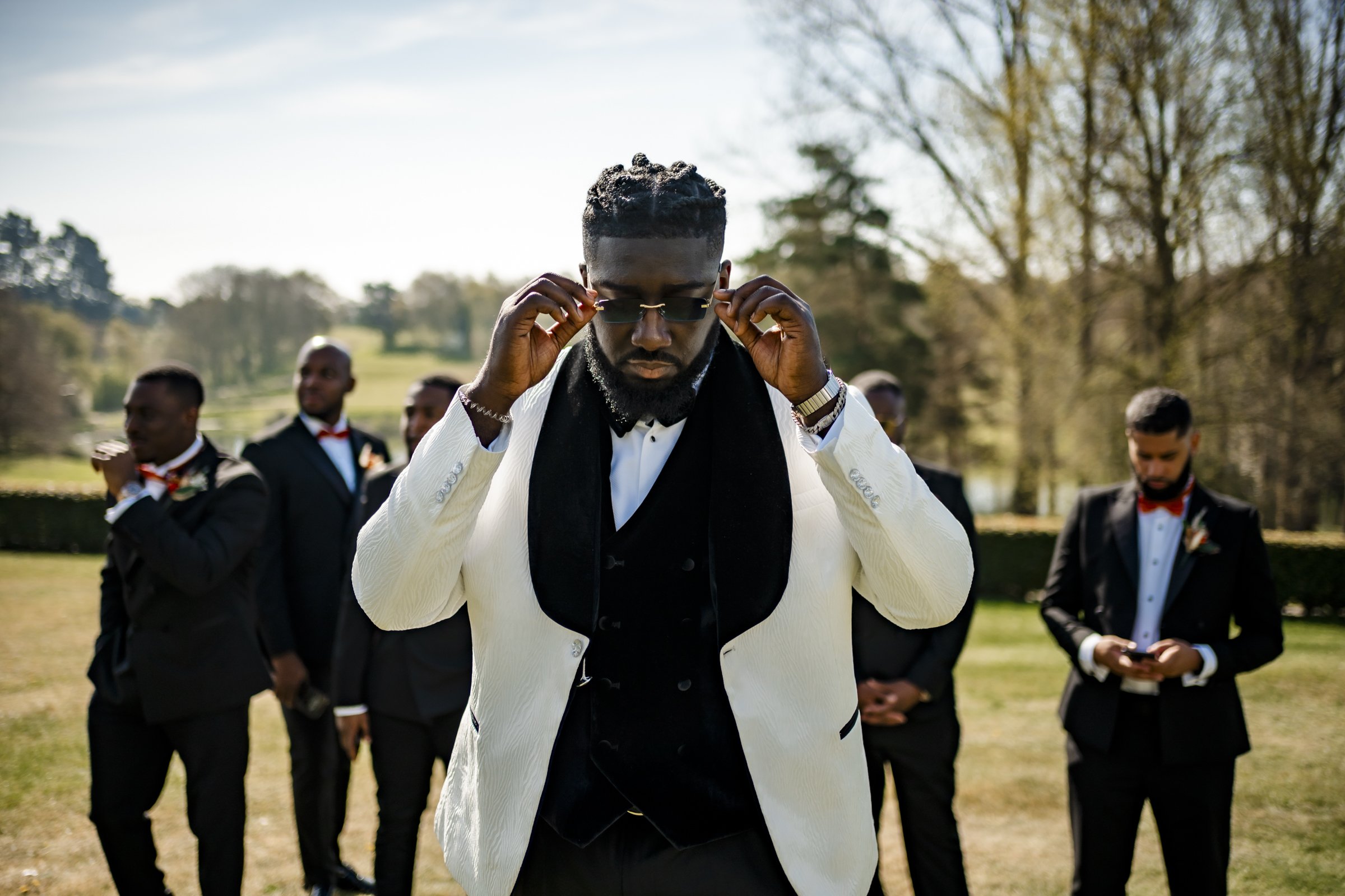 A man in a white tuxedo jacket and black vest adjusting sunglasses outdoors, with six men dressed in suits and bow ties in the background.