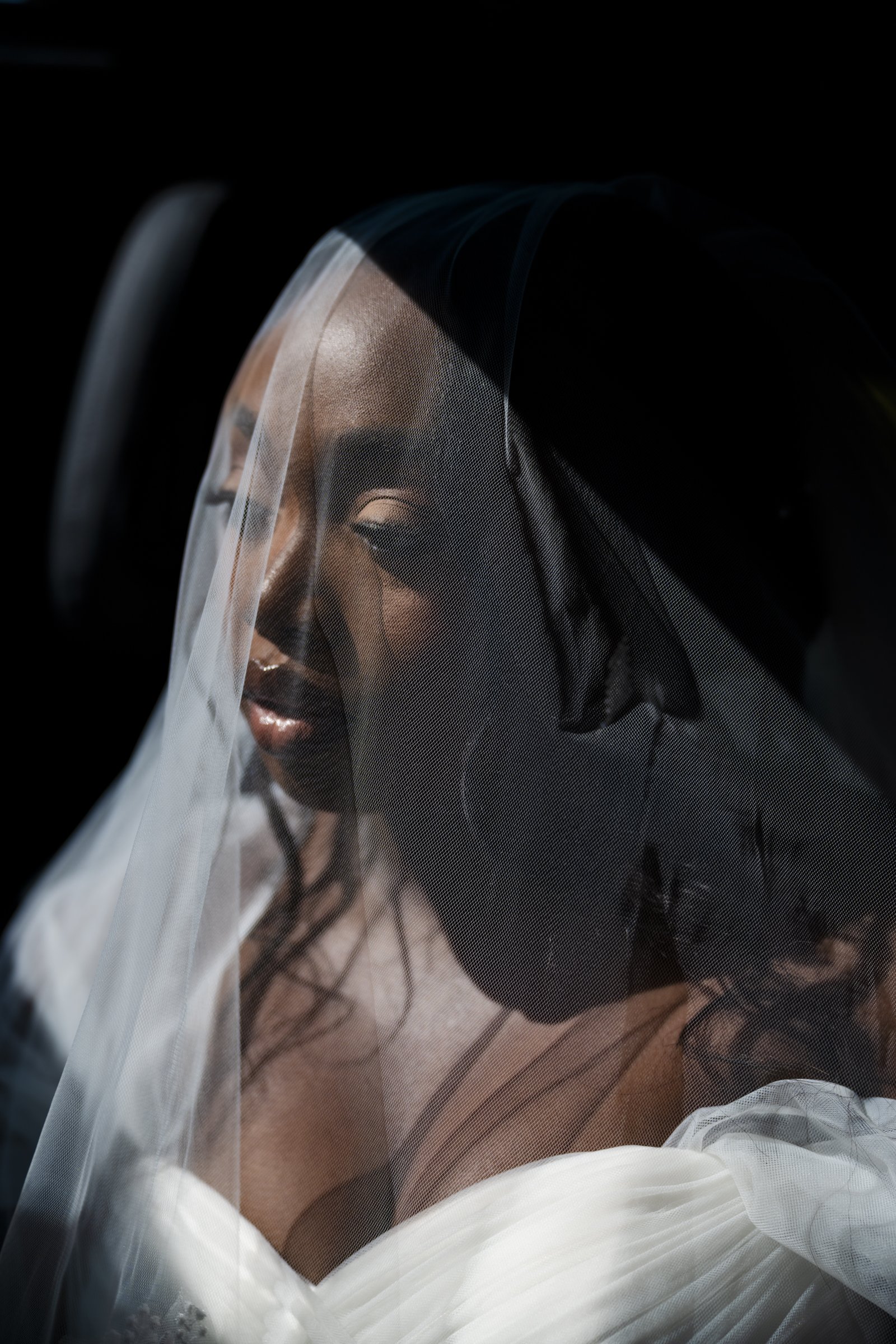 Ghanaian bride under a veil, wearing makeup including eye makeup and lipstick, dressed in a white gown, content before the Church wedding ceremony.
