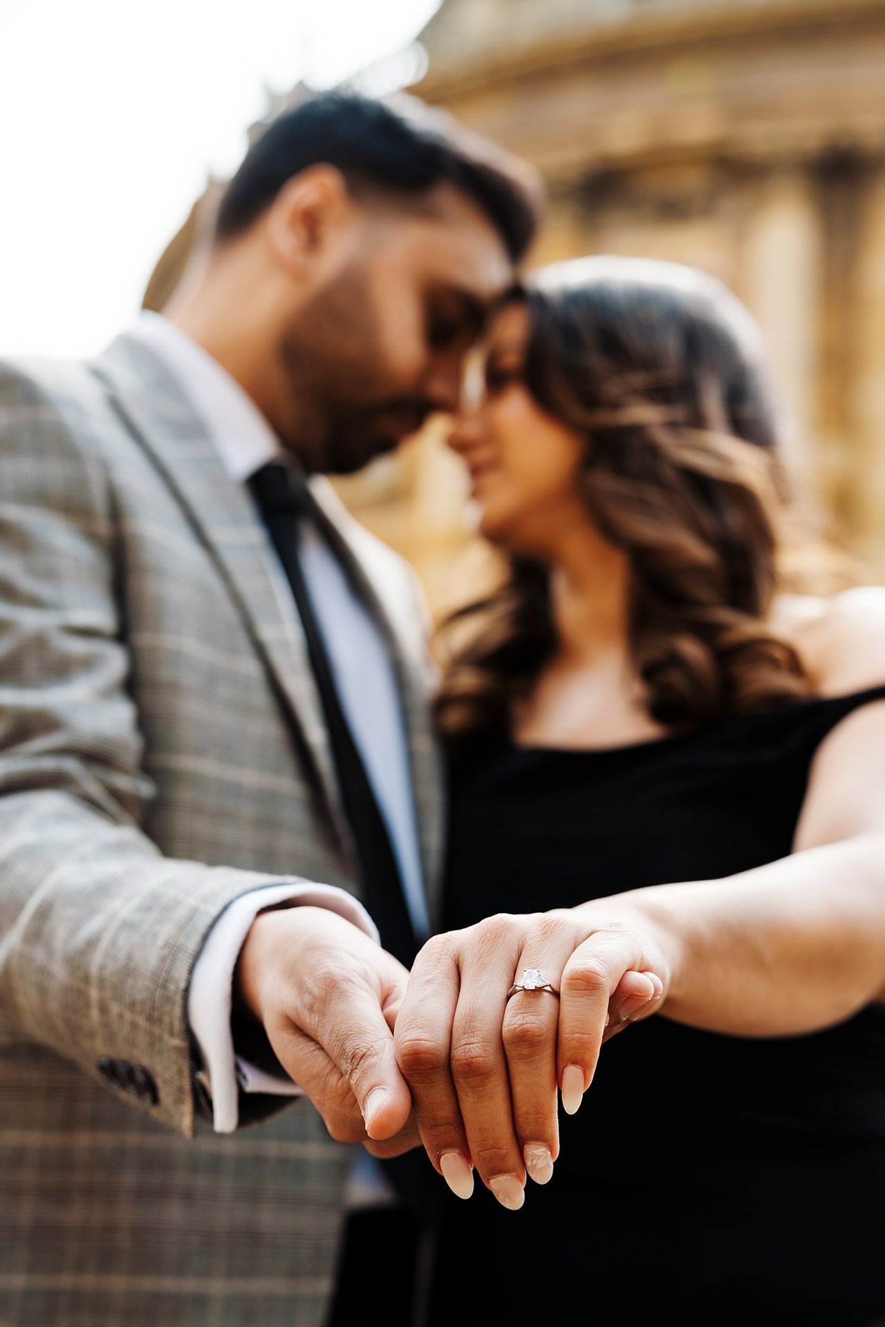 Close-up of a couple holding hands with an engagement ring, with blurred faces of a man and woman in romantic pose in the background.