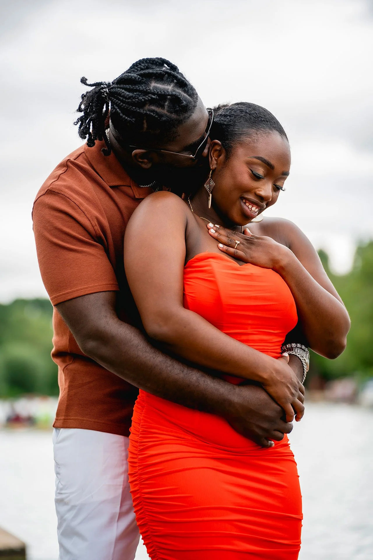 A couple embraces outdoors near a lake, with the man kissing the woman's cheek, the woman smiling, wearing a red dress, the man in a brown shirt and white pants.