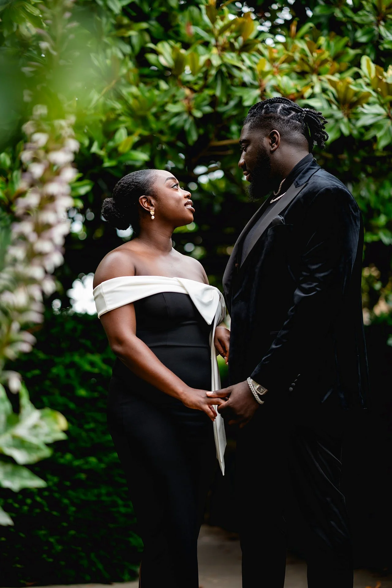 Ghanian couple dressed in formal attire holding hands and looking into each other's eyes outdoors, surrounded by lush greenery. Taken in Oxford.