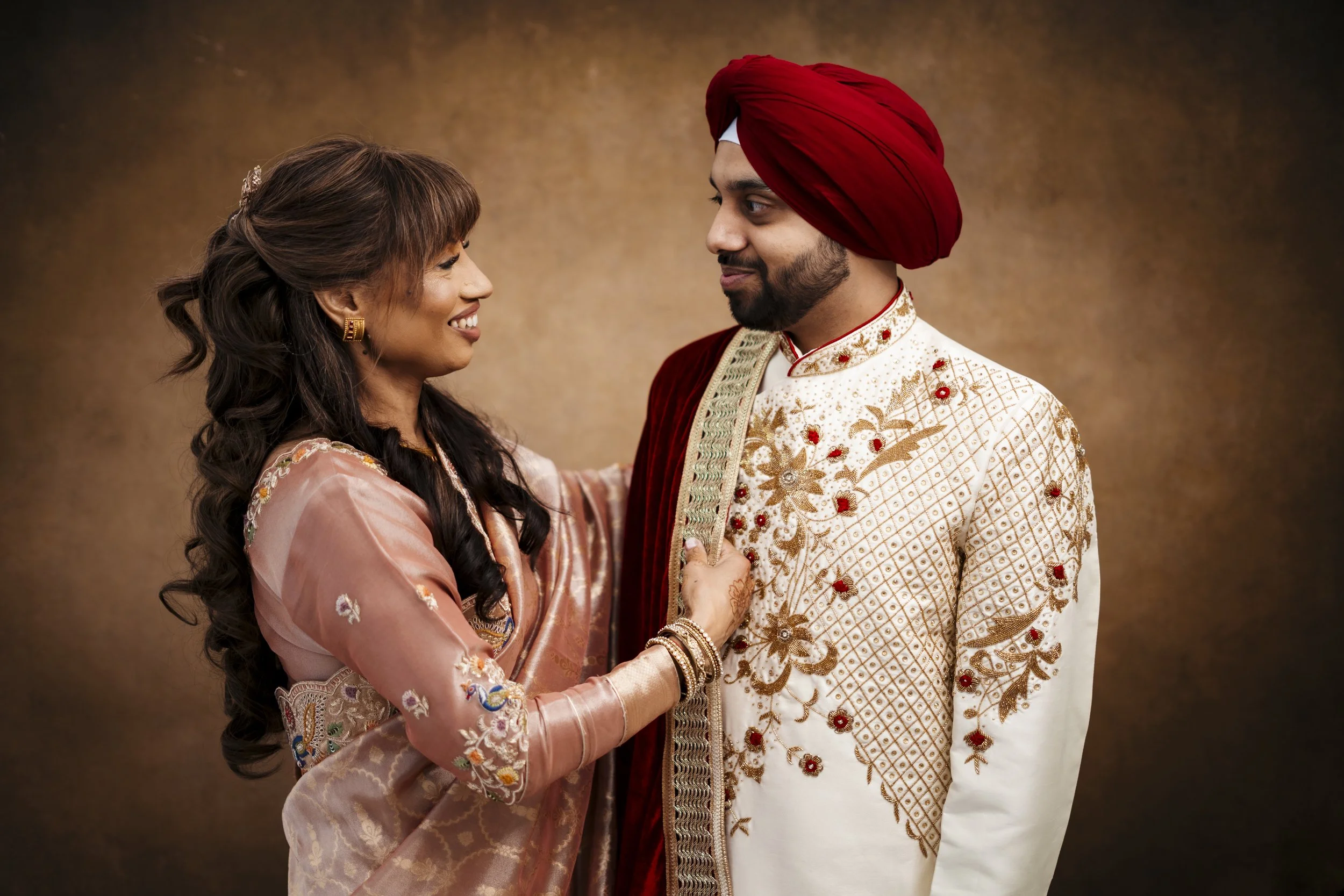 Indian couple dressed in traditional attire looking at each other, woman smiling and touching man’s outfit, man wearing a cream and gold embroidered sherwani with a red turban and shawl.