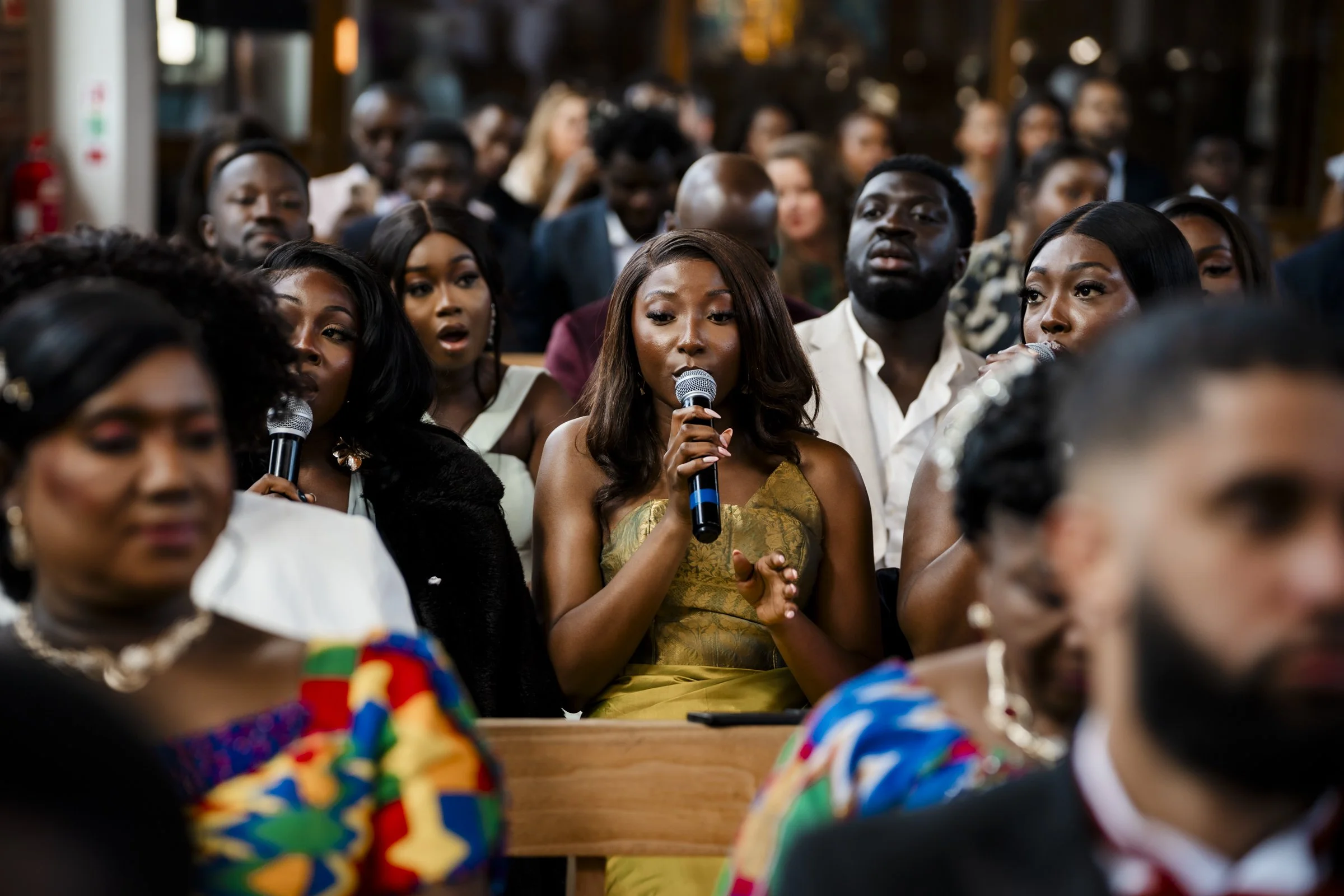 A woman in a gold dress speaking into a microphone at a crowded event with many guests seated in rows behind her.