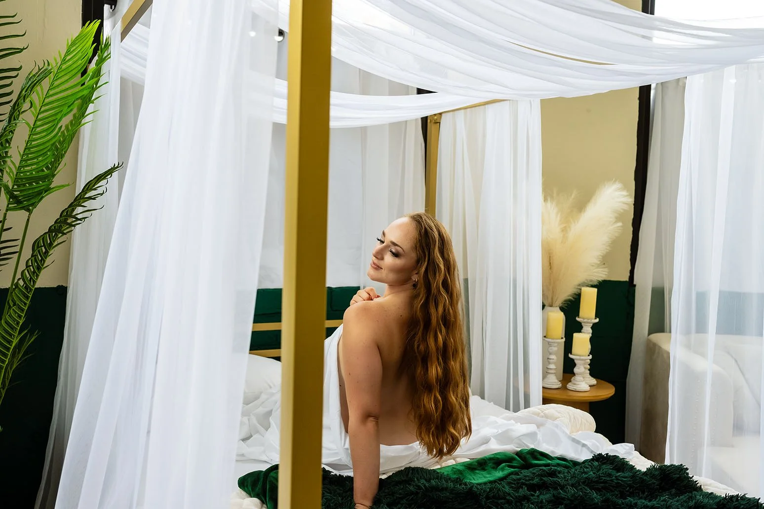 A woman with long, curly red hair sitting on a bed in a bedroom decorated with white curtains, candles, and greenery.