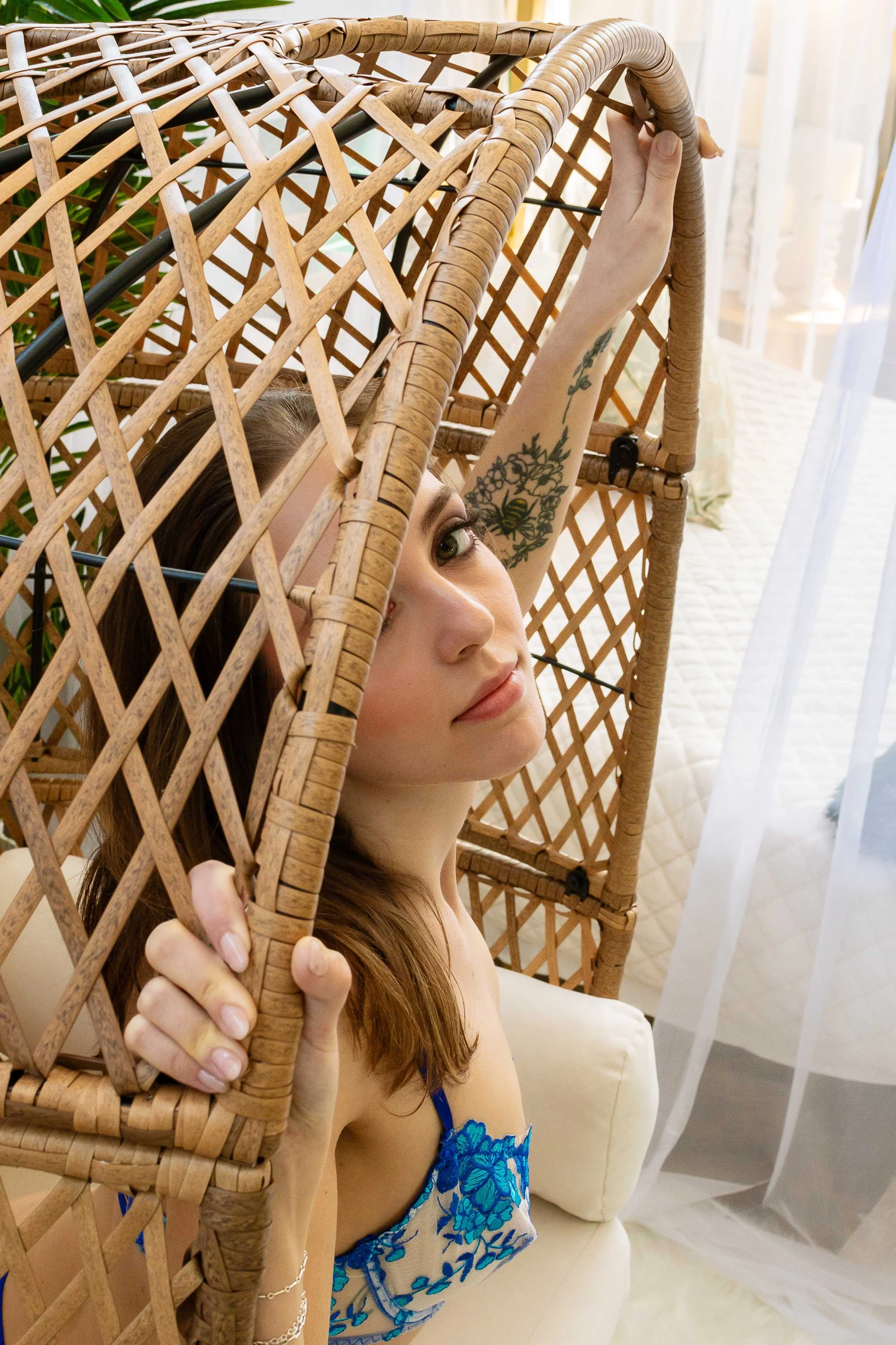 A woman with brown hair and tattoos holding a wicker privacy screen, peeking through the gaps, sitting on a cream-colored sofa.