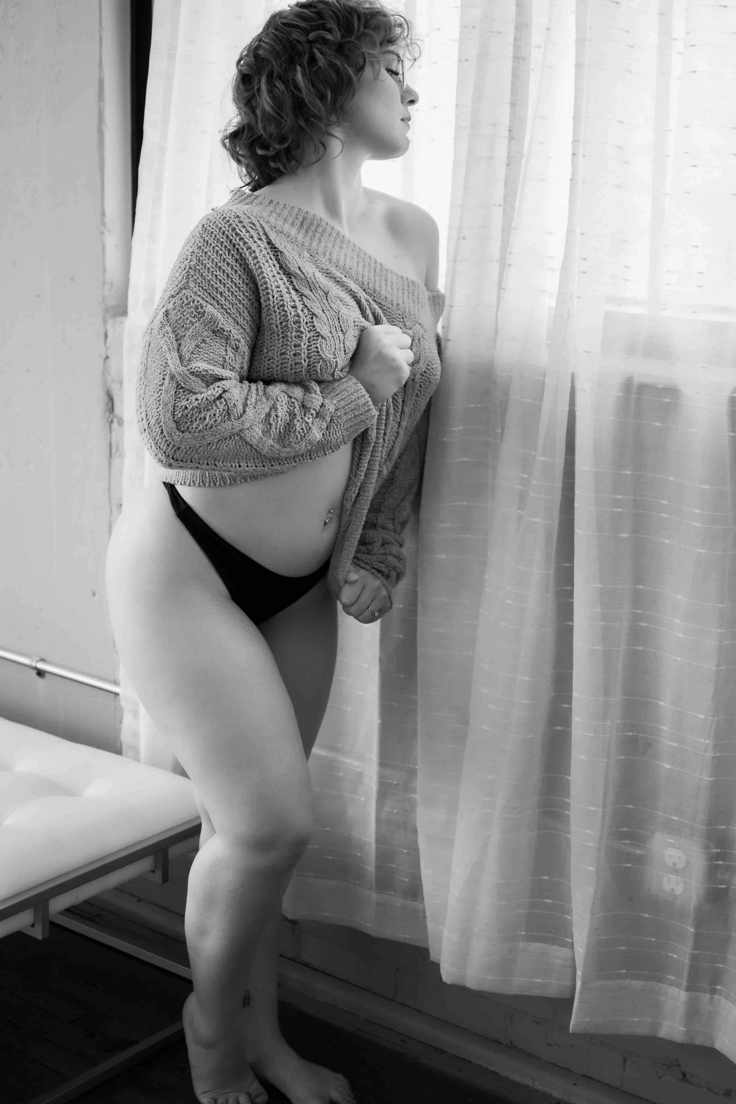 A woman stands near a window, partially undressed, wearing a sweater and black underwear, with a contemplative expression.