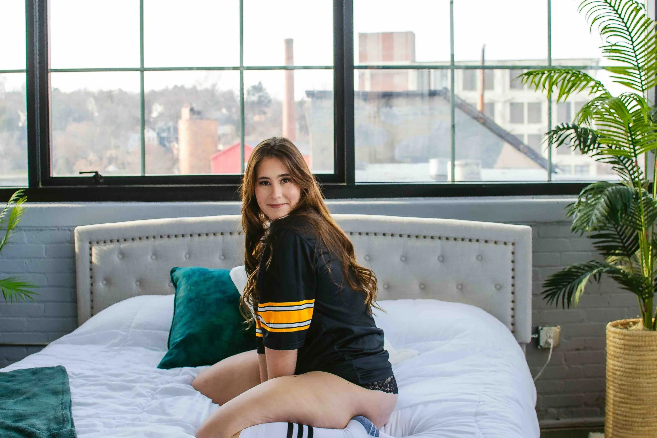 Young woman sitting on a bed in a bright bedroom with large windows, white brick wall, potted plant, and cityscape view outside.