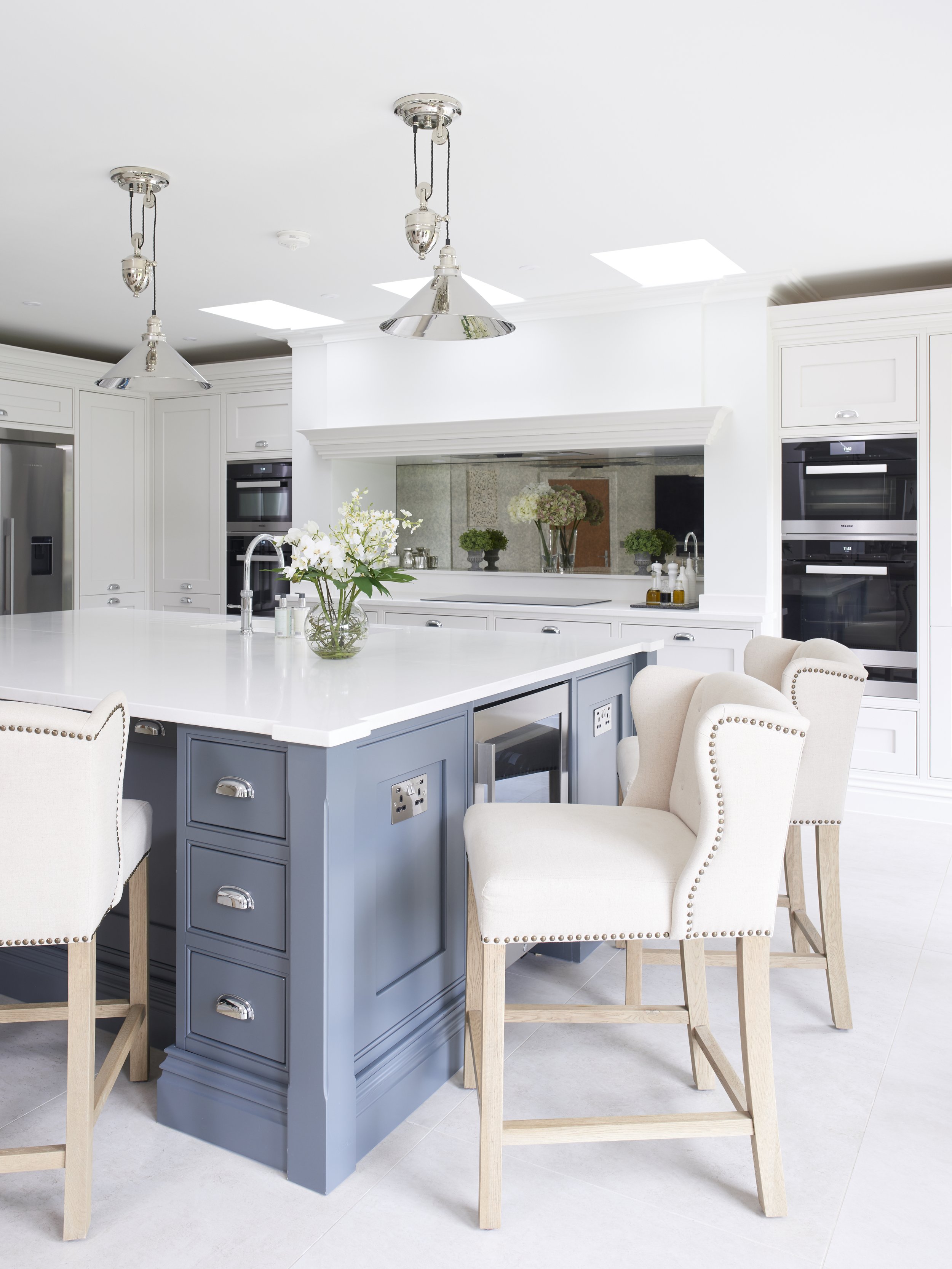 Modern kitchen with white cabinetry, a large kitchen island with gray-blue base and white countertop, beige upholstered bar stools, stainless steel appliances, pendant lighting, and decorative plants.
