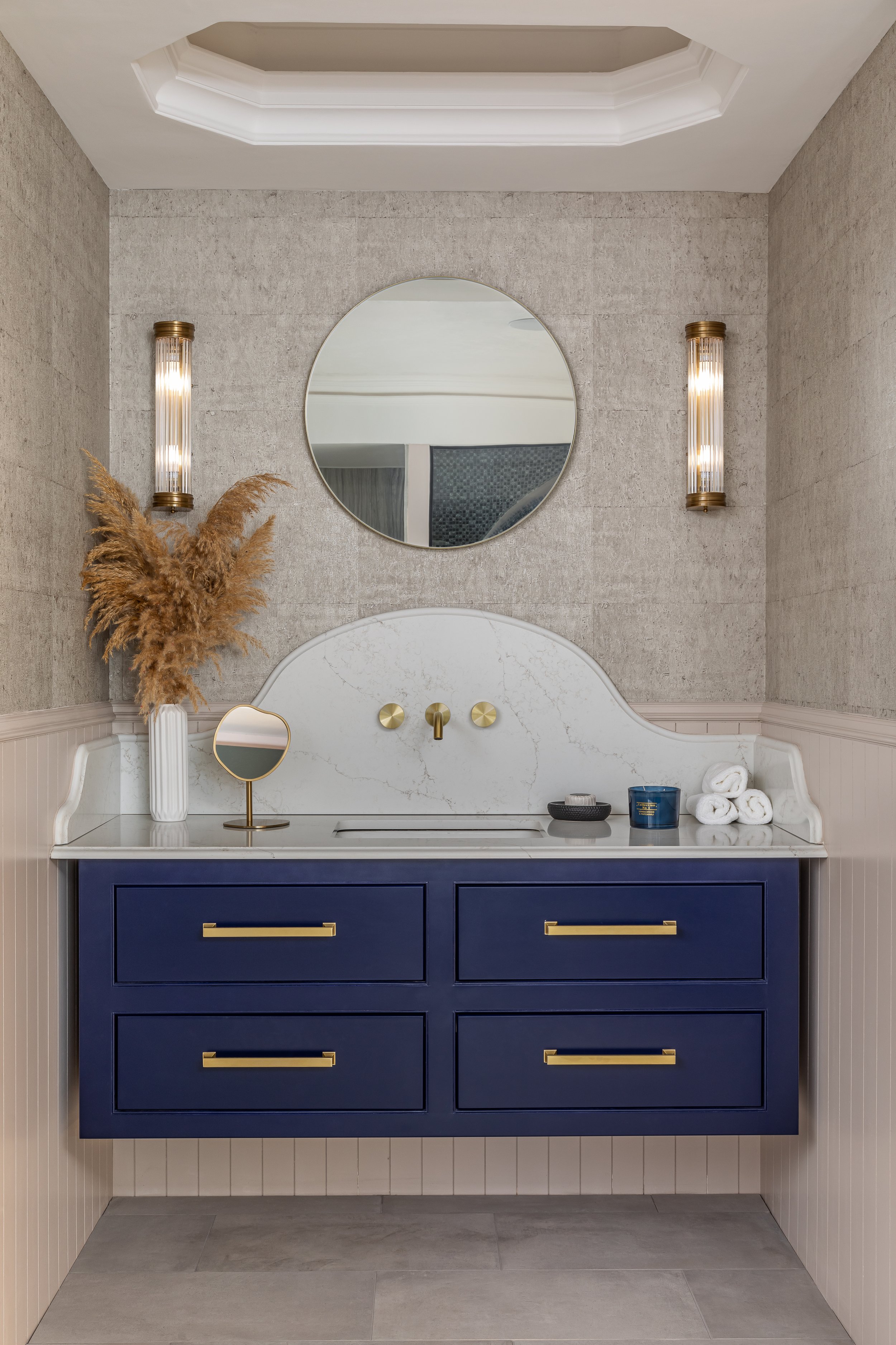 Elegant bathroom with a round mirror above a blue vanity, gold faucets, side wall sconces, and decorative items including dry pampas grass, candles, and rolled towels.