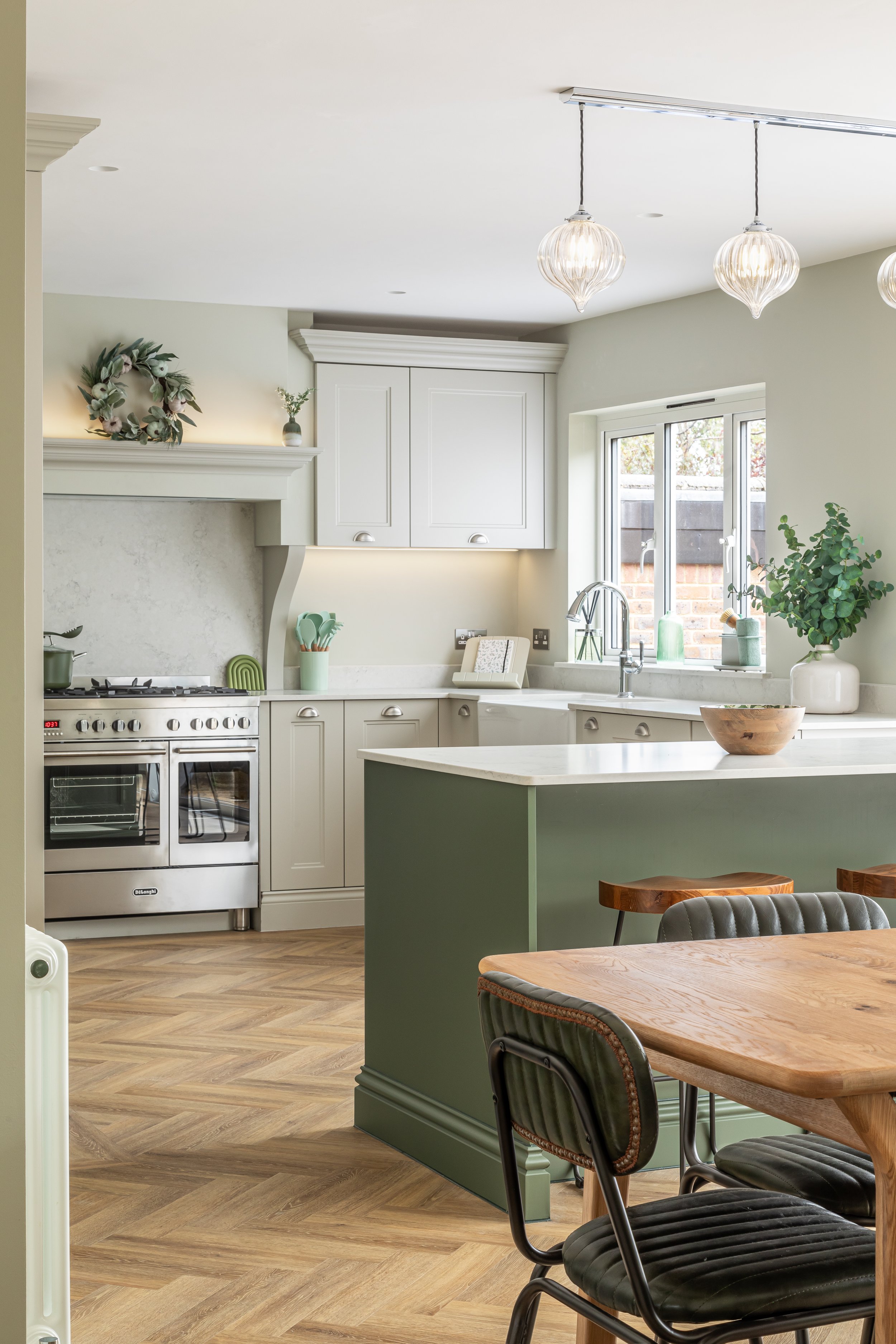Modern kitchen with light green cabinets, stainless steel stove, marble countertops, and wood flooring. A dining area with wooden table and chairs is visible, along with hanging pendant lights.