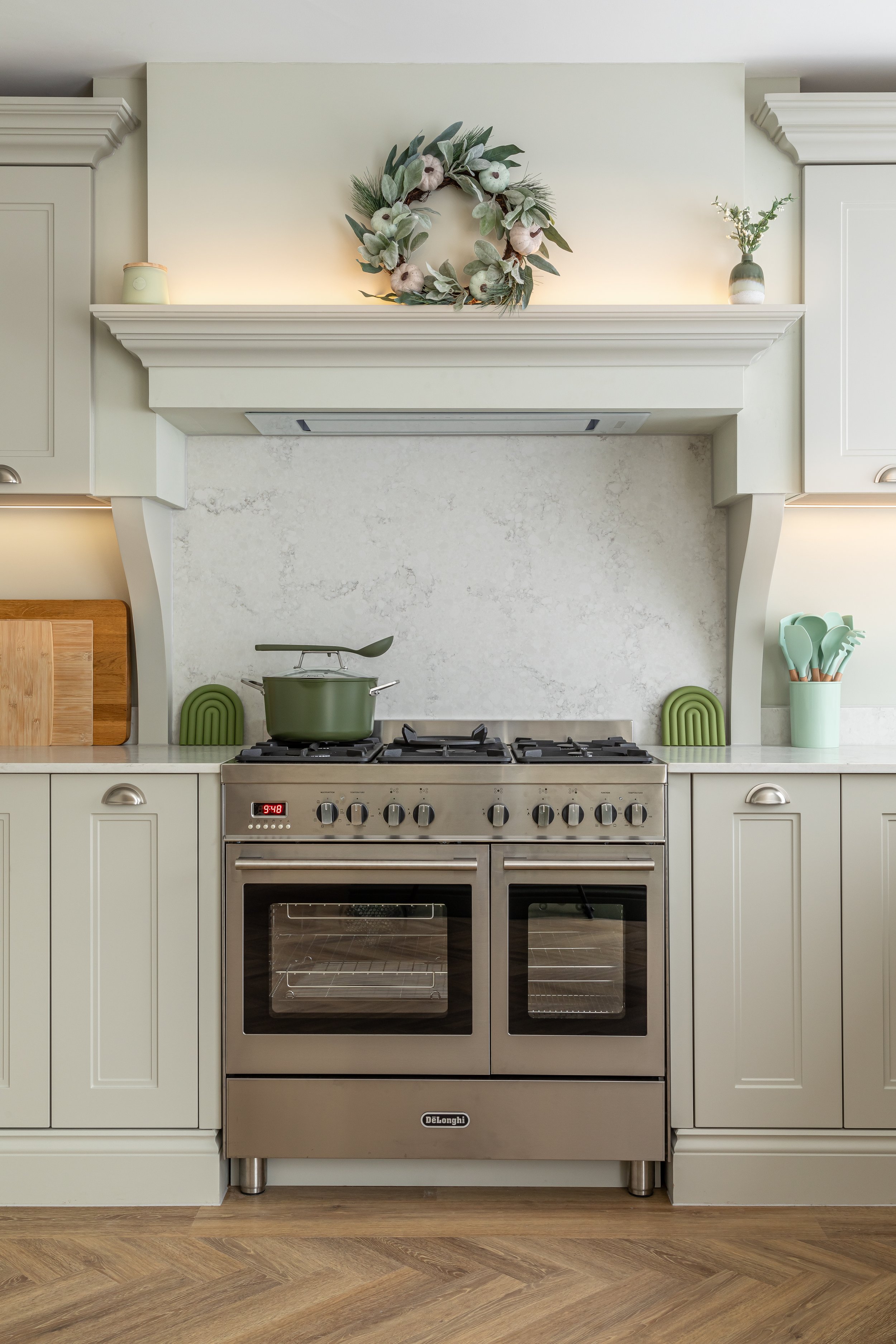 Modern kitchen with a stainless steel gas stove, olive green pot, decorative wreath, cutting boards, and utensils in light-colored cabinets.