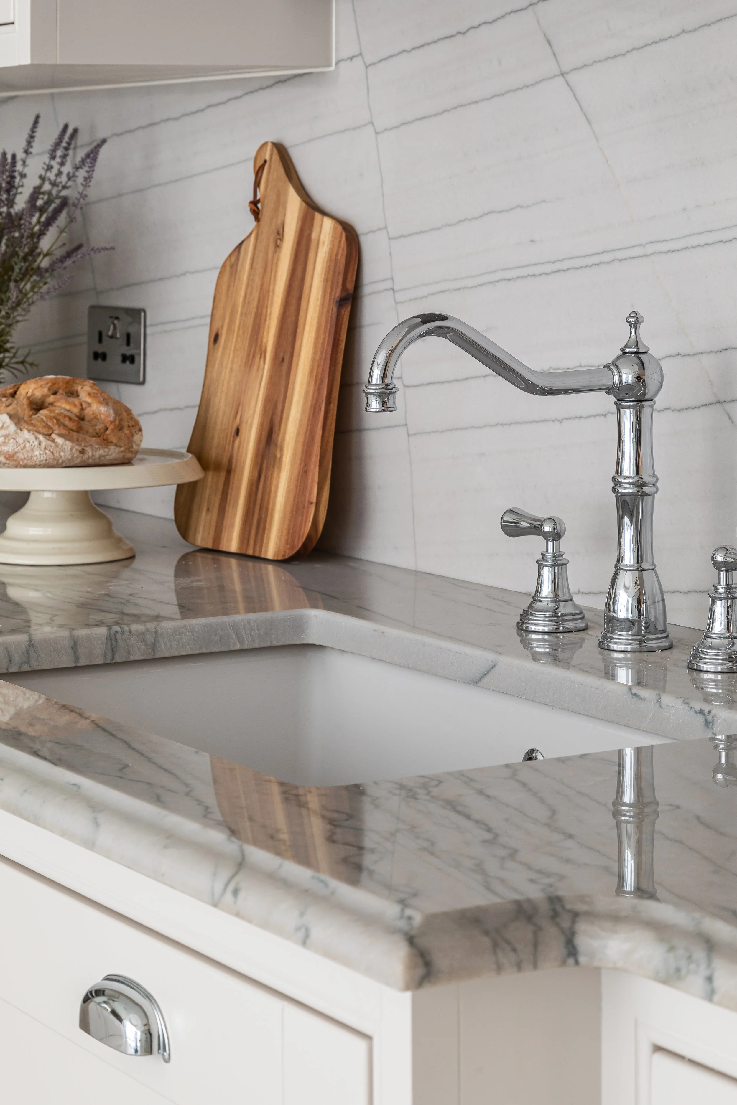 Marble kitchen countertop with a chrome faucet, wooden cutting board, bread on a stand, and lavender in the background.