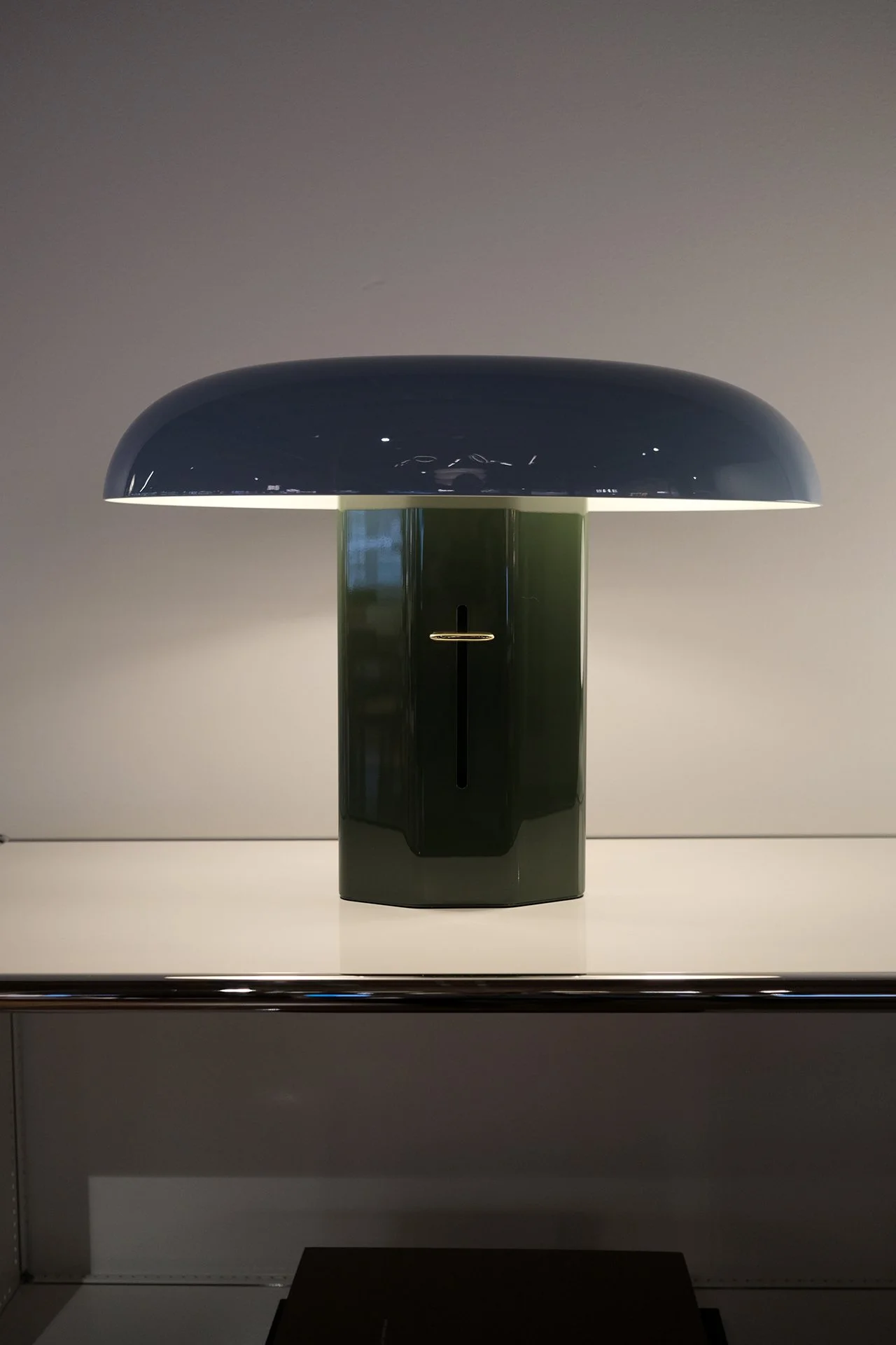 A modern table lamp with a dark blue, rounded shade and a green cylindrical base, placed on a white surface.