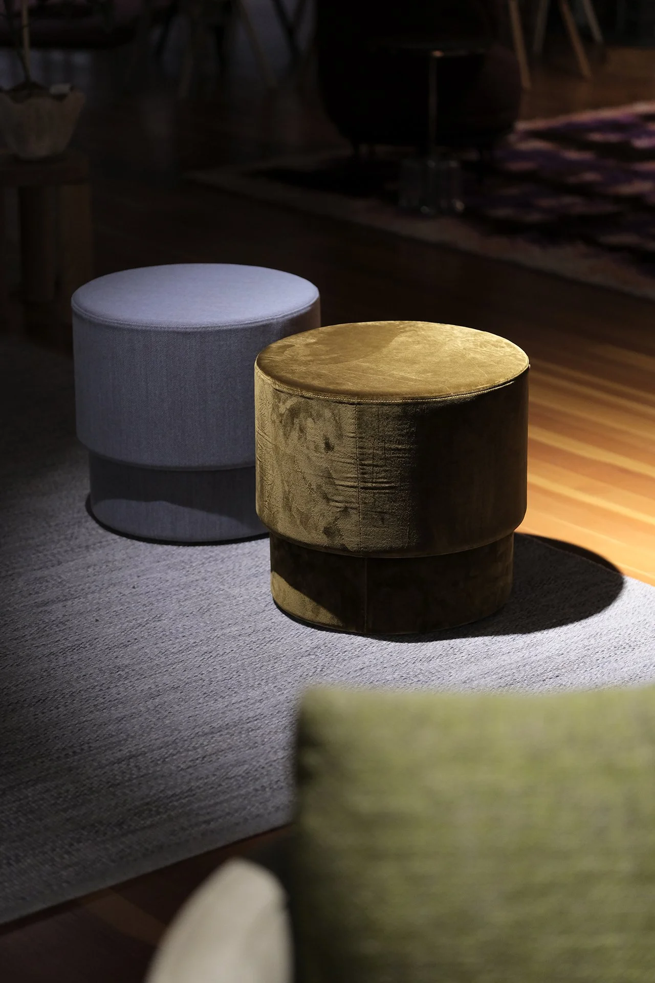 Two round ottomans, one light gray and one dark gold, placed on a carpet in a dimly lit room.
