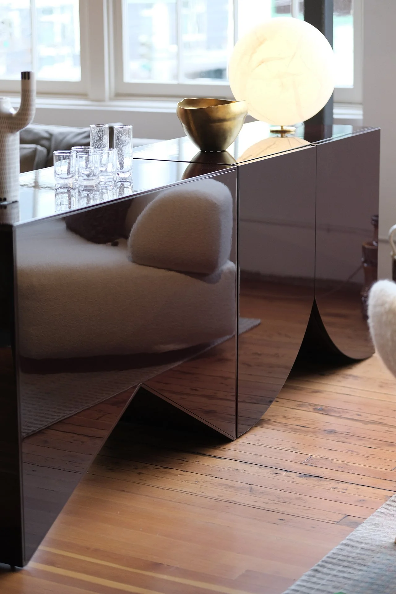 A black, reflective, curved sideboard with decorative items on top, including a glowing lamp, a gold bowl, and several glasses, with a dining table and windows in the background.