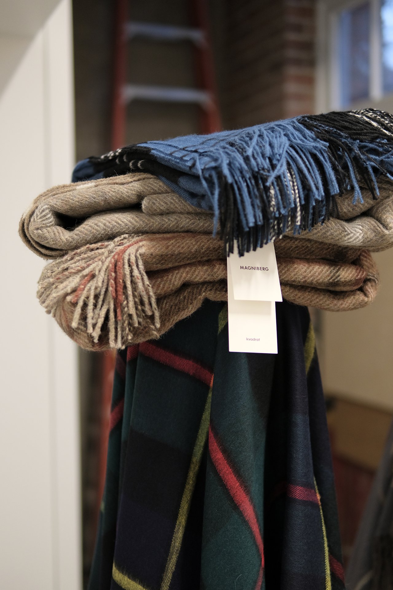 A stack of folded scarves on a black stand, with a brick wall, a window, and a ladder in the background.